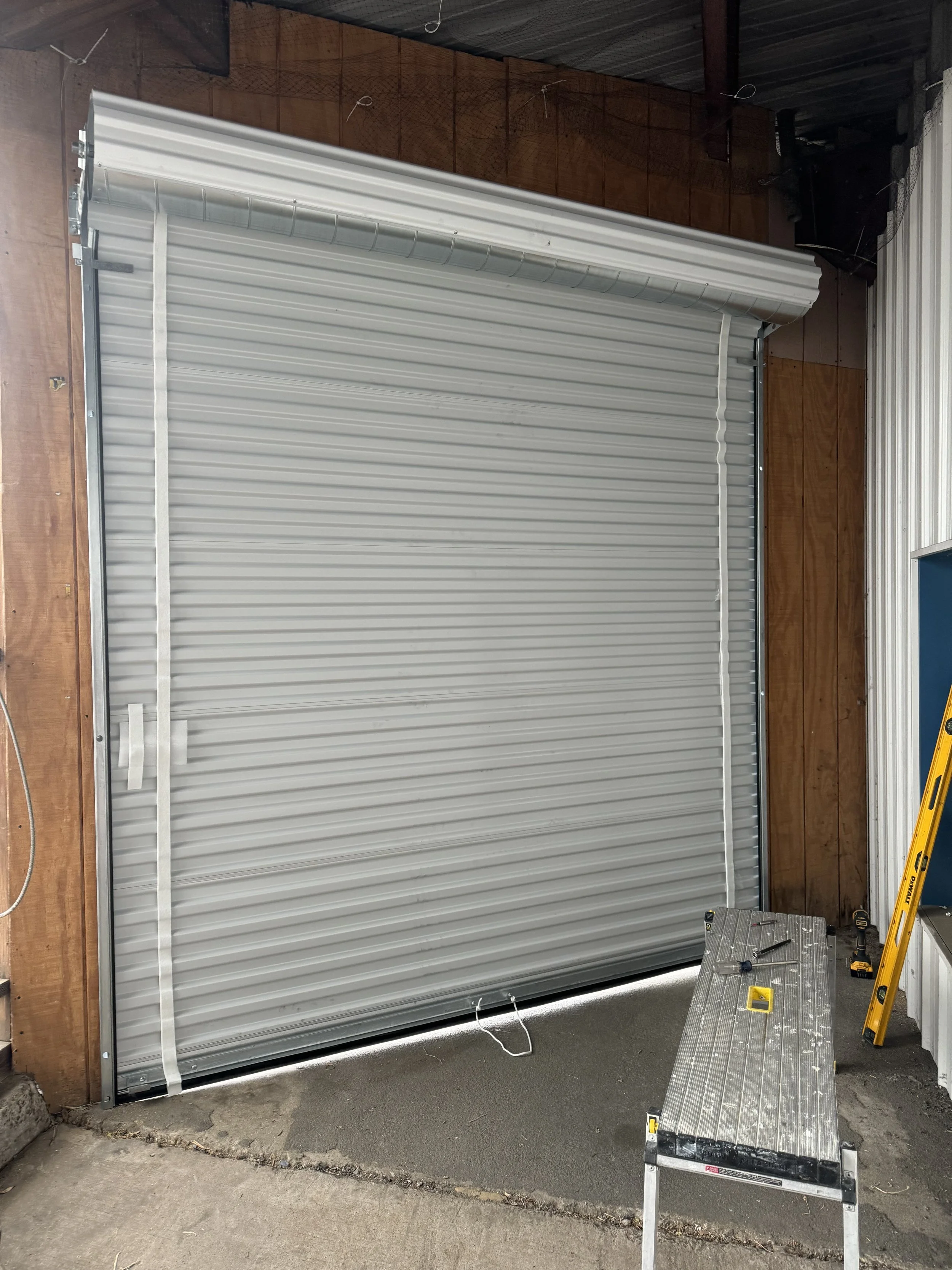 Newly installed white metal garage door partially open, with tools and ladder nearby, in a garage with wooden walls.