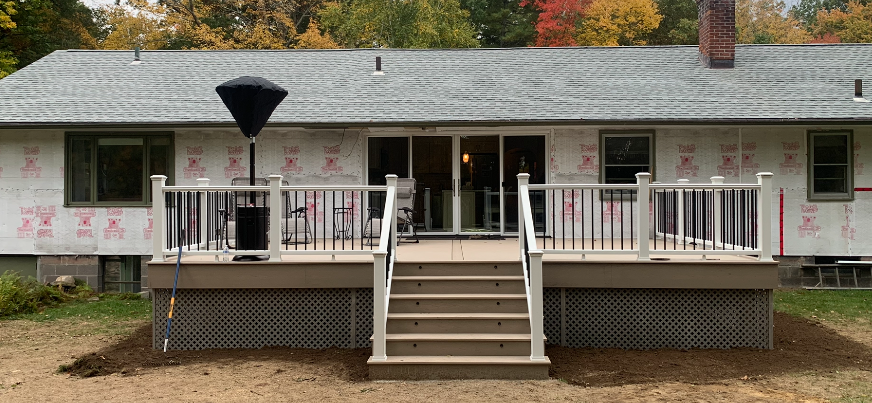 Backyard deck under construction on a house with a gray roof, black and white railing, steps leading down to a dirt yard, and a large patio umbrella.