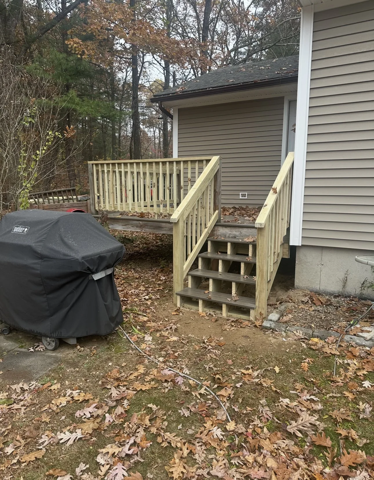 A wooden staircase with handrails leading up to a small raised deck attached to the back of a house with beige vinyl siding. Fallen leaves cover the ground, and a black grill with a cover is in the foreground. Trees with autumn foliage are in the bac