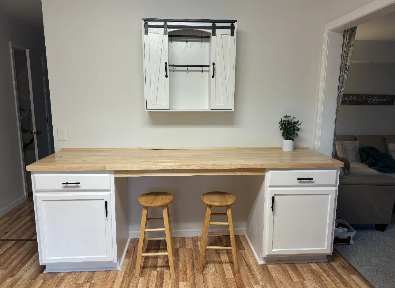 Kitchen bar area with two wooden stools, a light wood countertop, white cabinets, and a small potted plant, with a wall-mounted cabinet featuring black hardware, and a living room in the background.