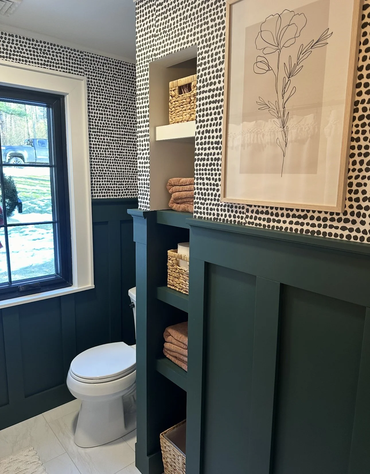 Bathroom with a window, teal wainscoting, shelves with baskets and folded towels, and a framed floral line drawing.