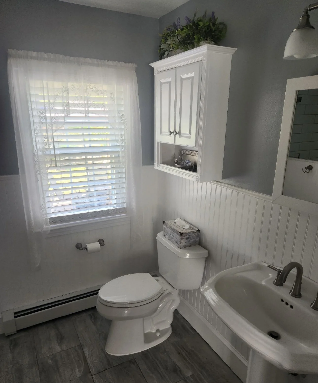 The image shows a small bathroom with a window covered with white lace curtains, a white toilet with a wooden tissue box on top, a white wall-mounted cabinet with a floral arrangement on top, a white sink with a silver faucet, and a mirror with a lig