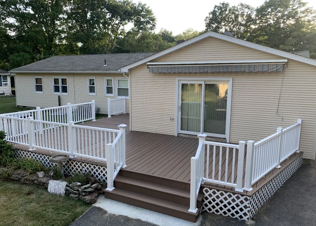 Backyard deck with white railing and steps, attached to a beige house with sliding glass door, striped awning, and multiple windows, surrounded by green trees.