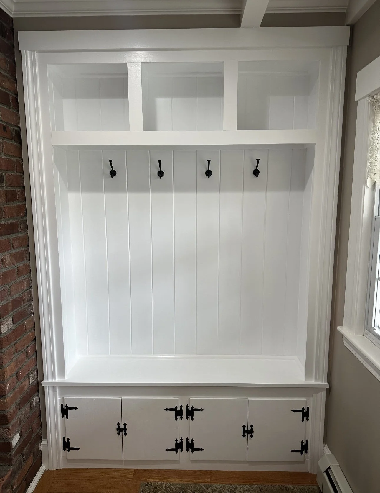 White built-in cabinet with upper glass display shelves, a middle section with four black hooks, and lower cabinets with black hardware, situated against a wall near a window.