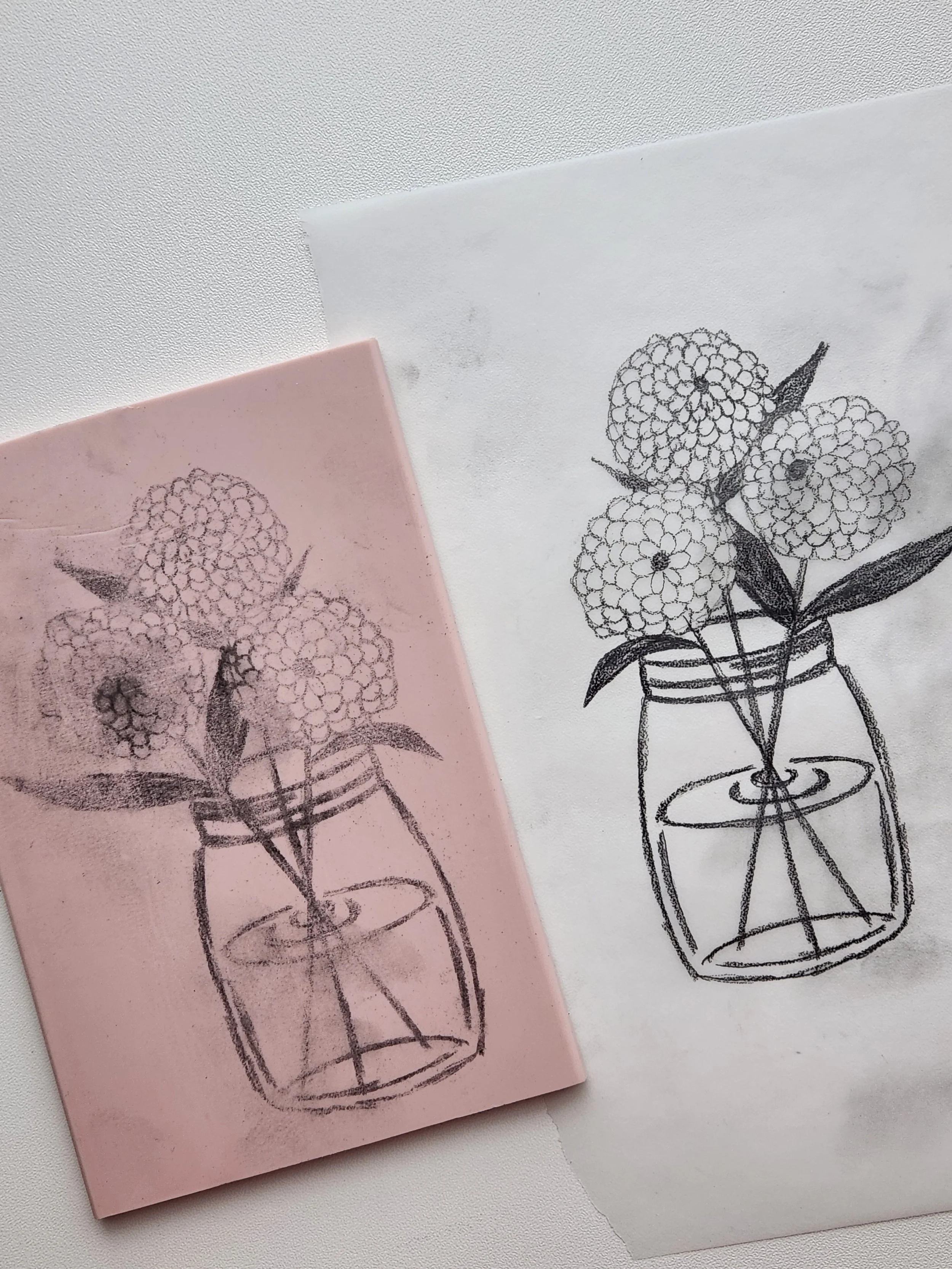 A black ink sketch of a bouquet of flowers in a round vase on a piece of paper and a pink sketchbook with the same drawing.