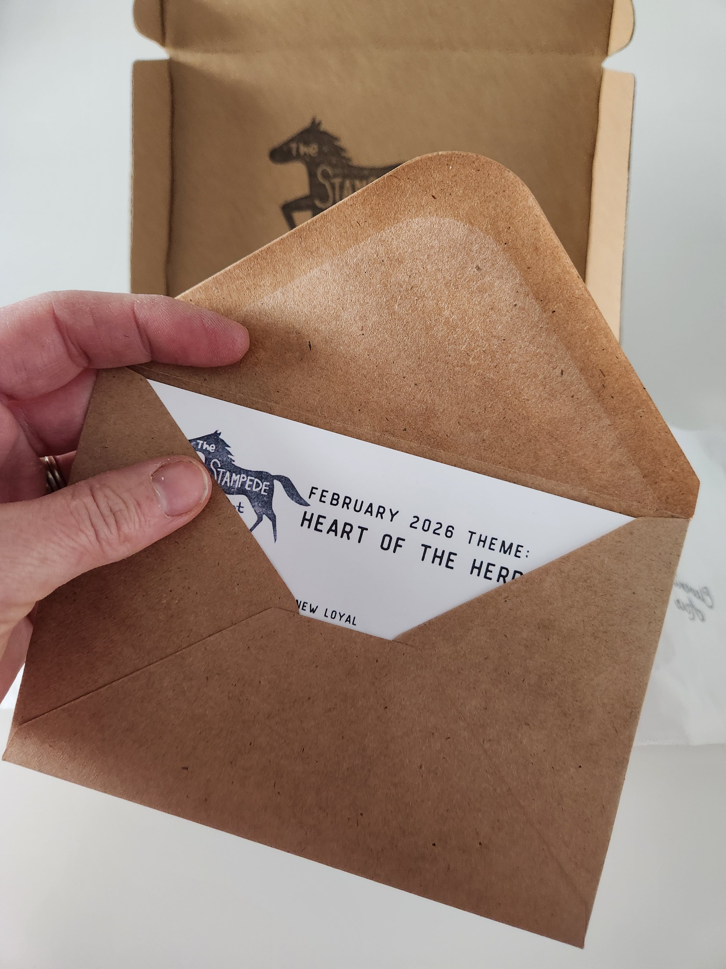 A hand holding a brown paper envelope with a white card inside, partially visible, with text about a February 2026 theme, 'Heart of the Hero', and a graphic of a horse. In the background, there's a paper bag with a black horse silhouette and the text 'The Stampede'.