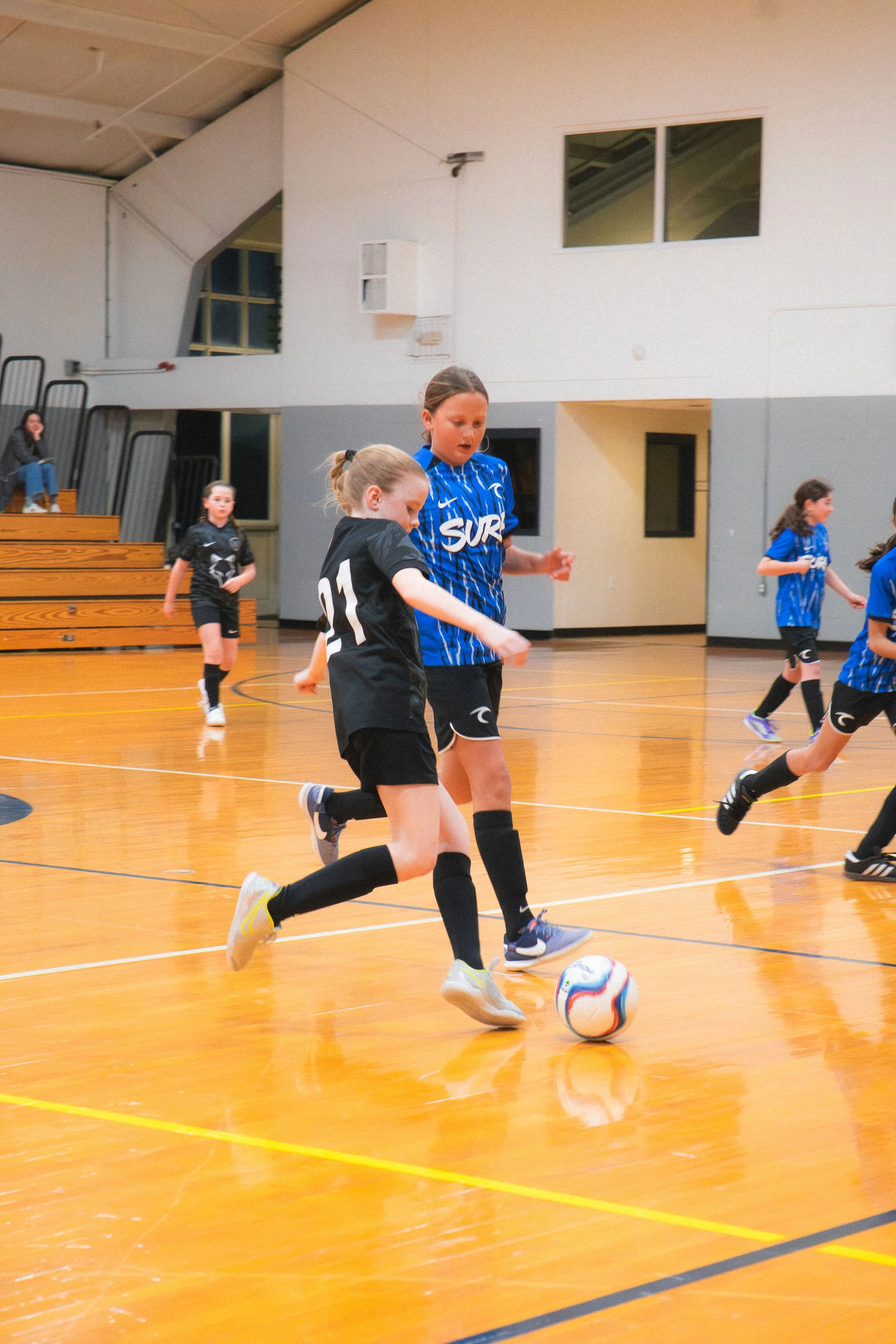 Young girls playing indoor soccer on a polished wooden court, some chasing the ball, in a gymnasium with bleachers in the background.