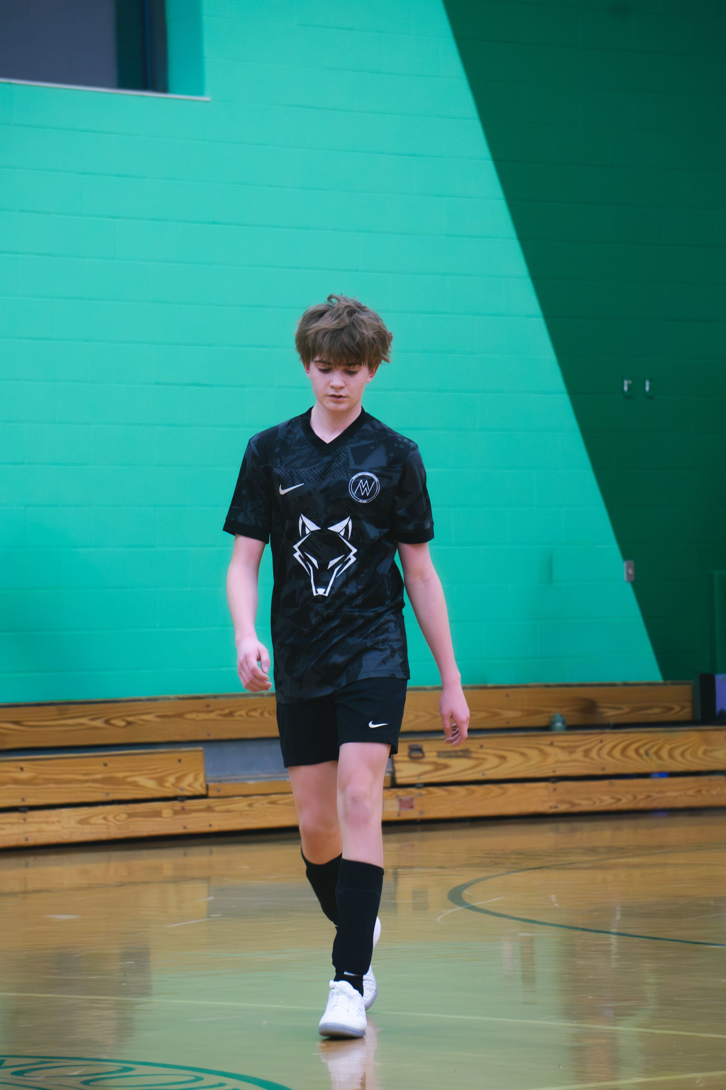 A young boy walking on an indoor basketball court, wearing a black sports uniform with a wolf logo on the shirt and Nike logo, and white sneakers.