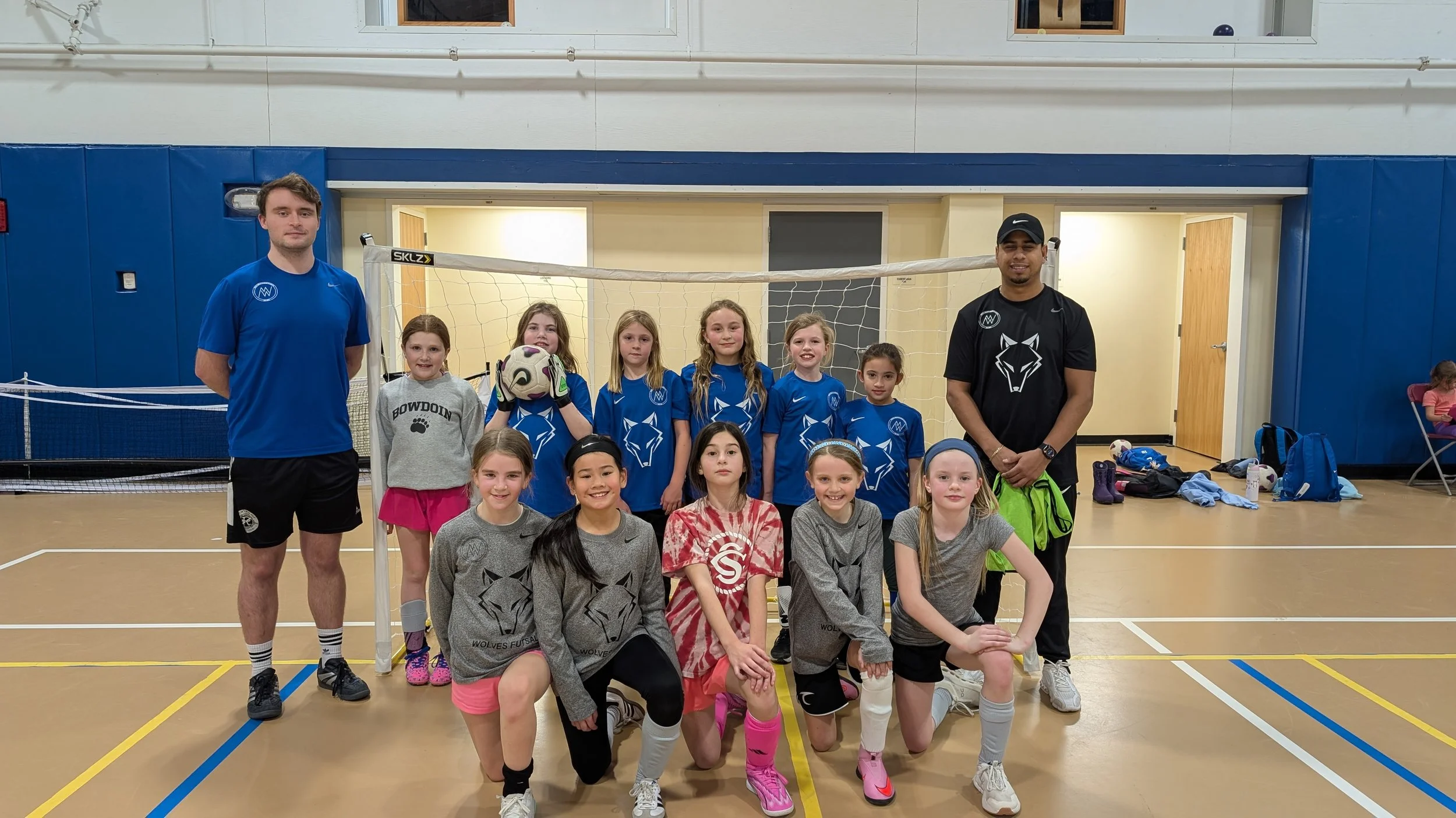 A girls' volleyball team with two coaches is posing for a photo in an indoor gymnasium, standing in front of a volleyball net.