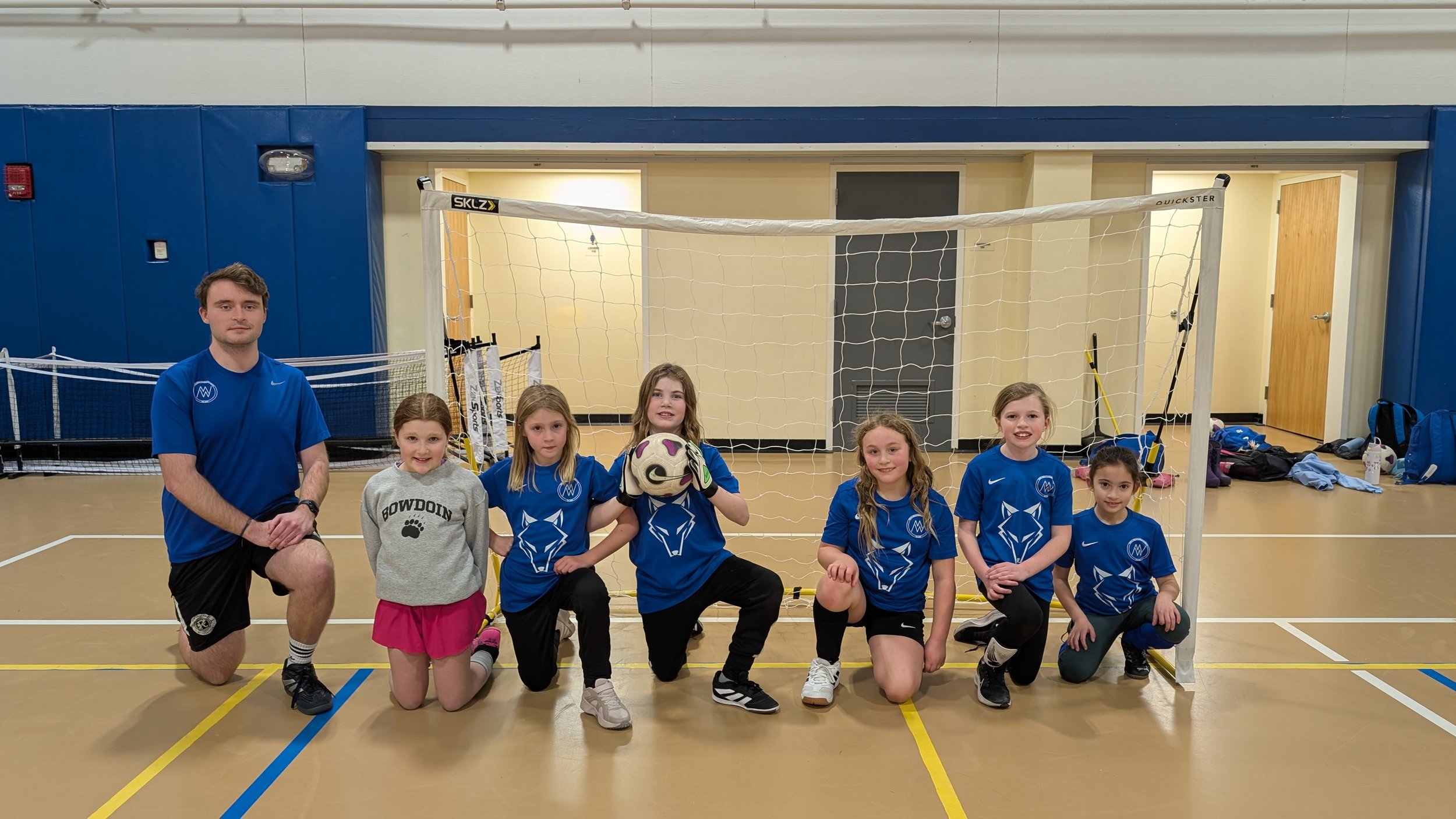 A youth indoor soccer team with six girls and a coach, kneeling and standing in front of a soccer goal on the gym floor. The girls wear matching blue jerseys with a wolf logo, black shorts, and various athletic shoes. One girl is holding a soccer bal