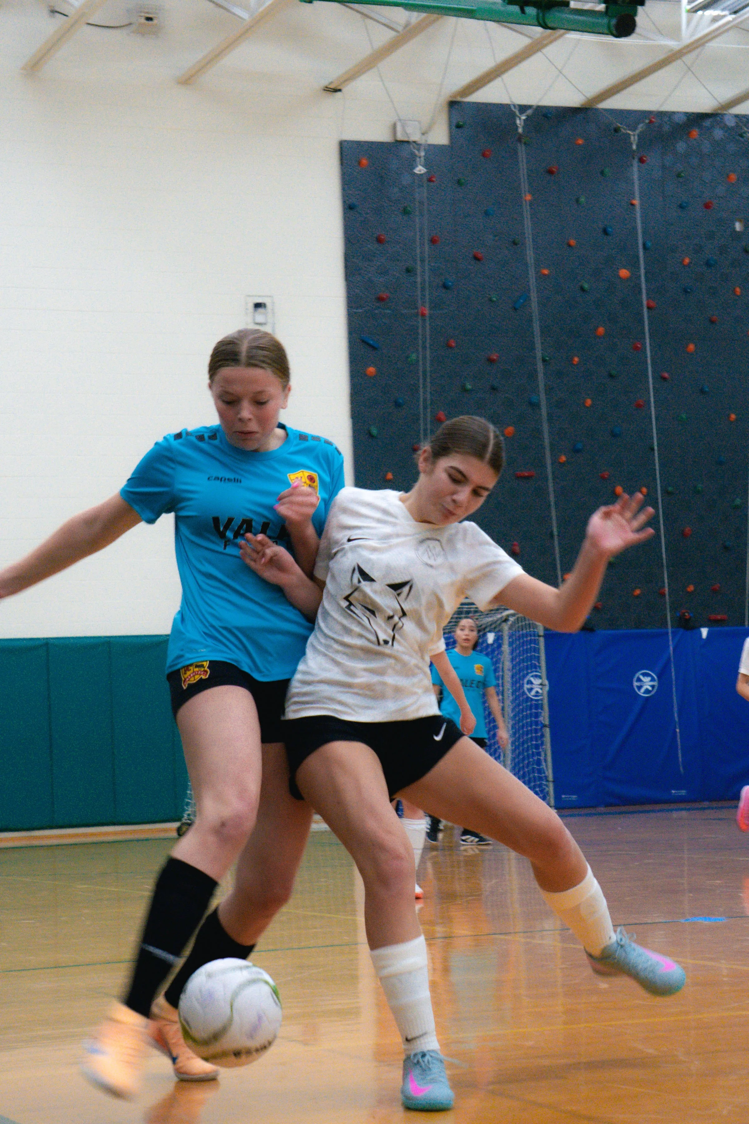 Two young women playing indoor soccer, fighting for control of the ball. One is wearing a blue jersey and the other a white jersey with a wolf design, on a gymnasium floor, with a climbing wall in the background.
