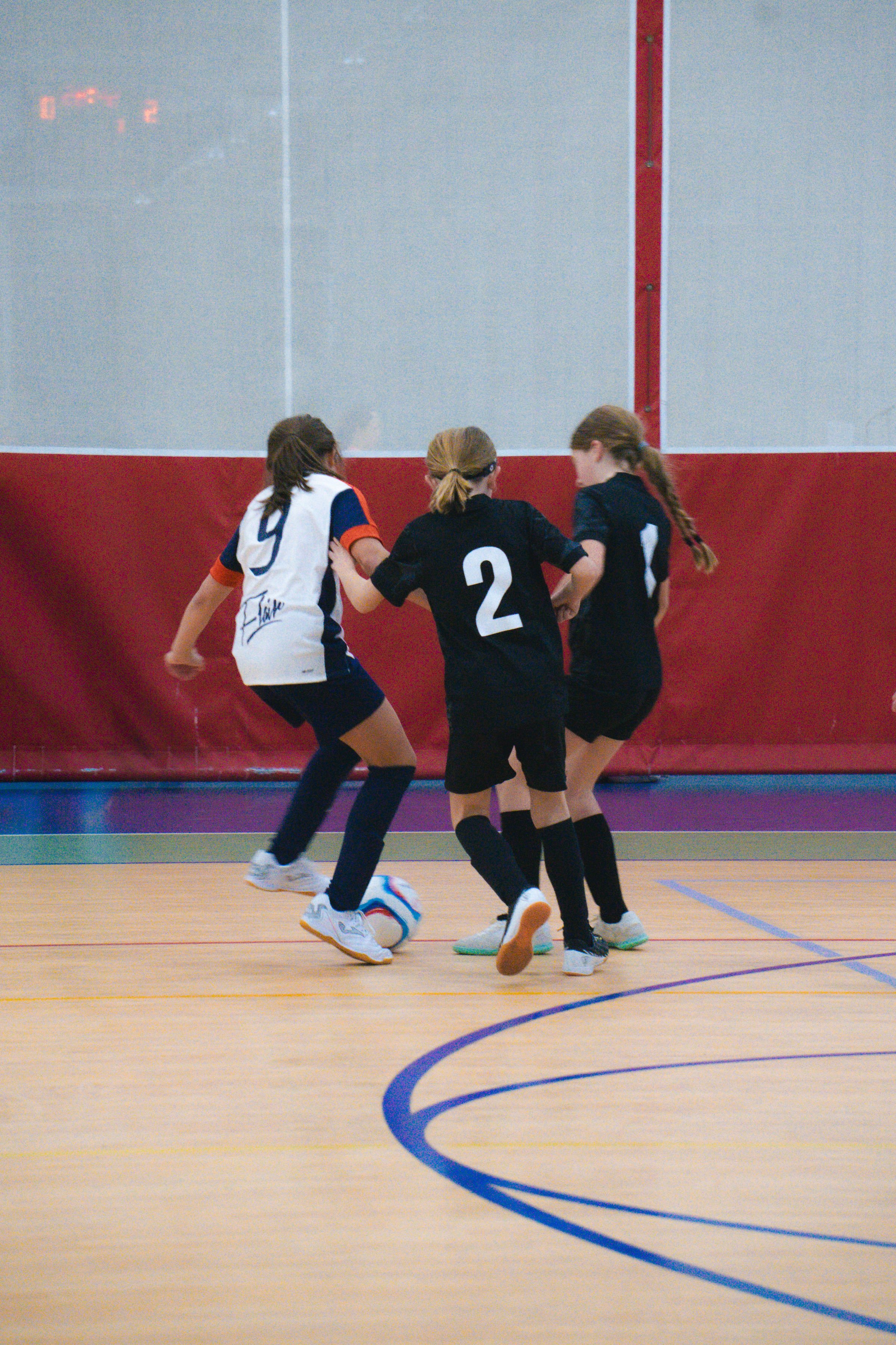 Three young girls playing indoor soccer on a court with a wooden floor, wearing black and white jerseys, two with long hair in braids, one with loose hair, all focused on the soccer ball.