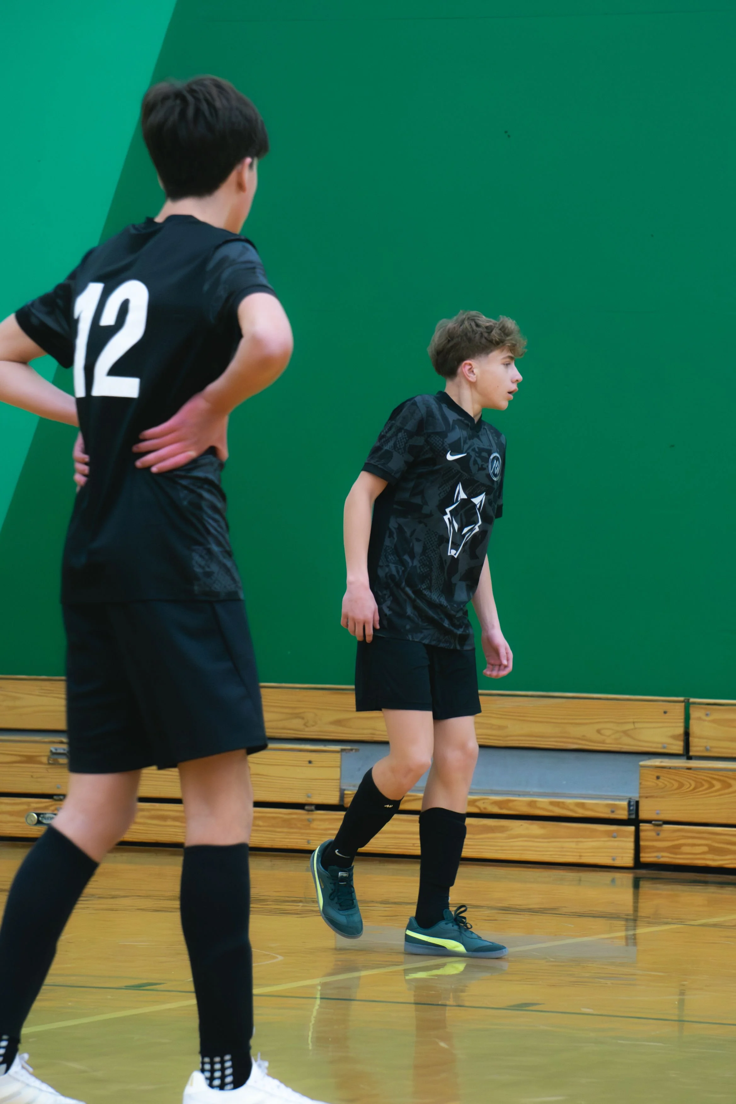 Two young boys playing indoor soccer on a gym court with green walls. One boy is seen from the back, wearing a black jersey with the number 12, and the other is facing sideways, wearing a black sports jersey and black shorts.
