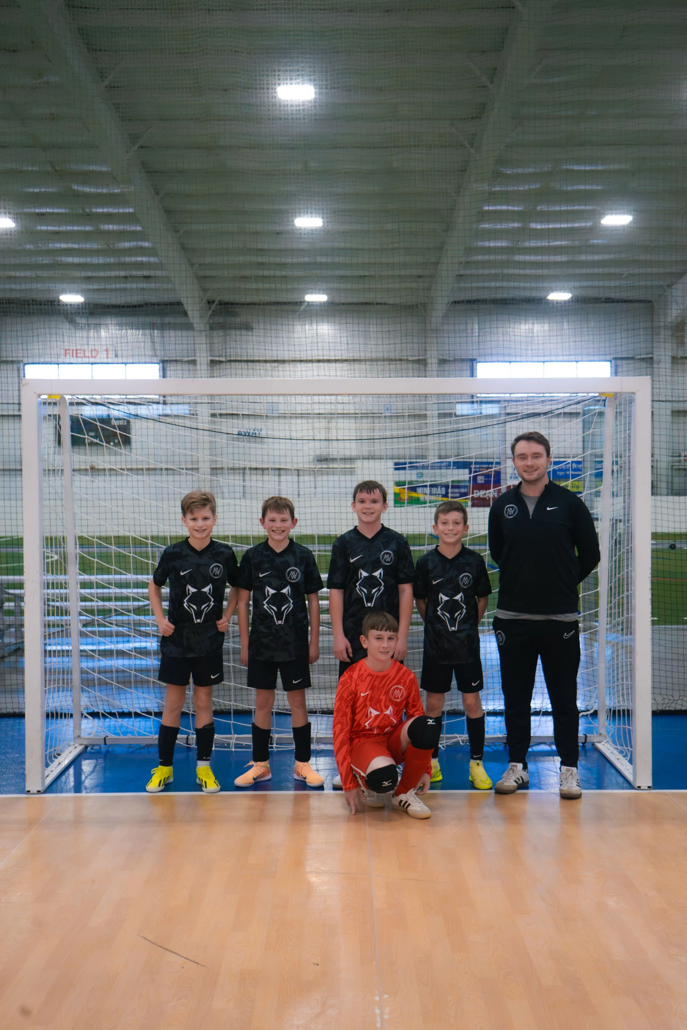 Group of young soccer players and coach posing in front of an indoor soccer goal in a sports facility.