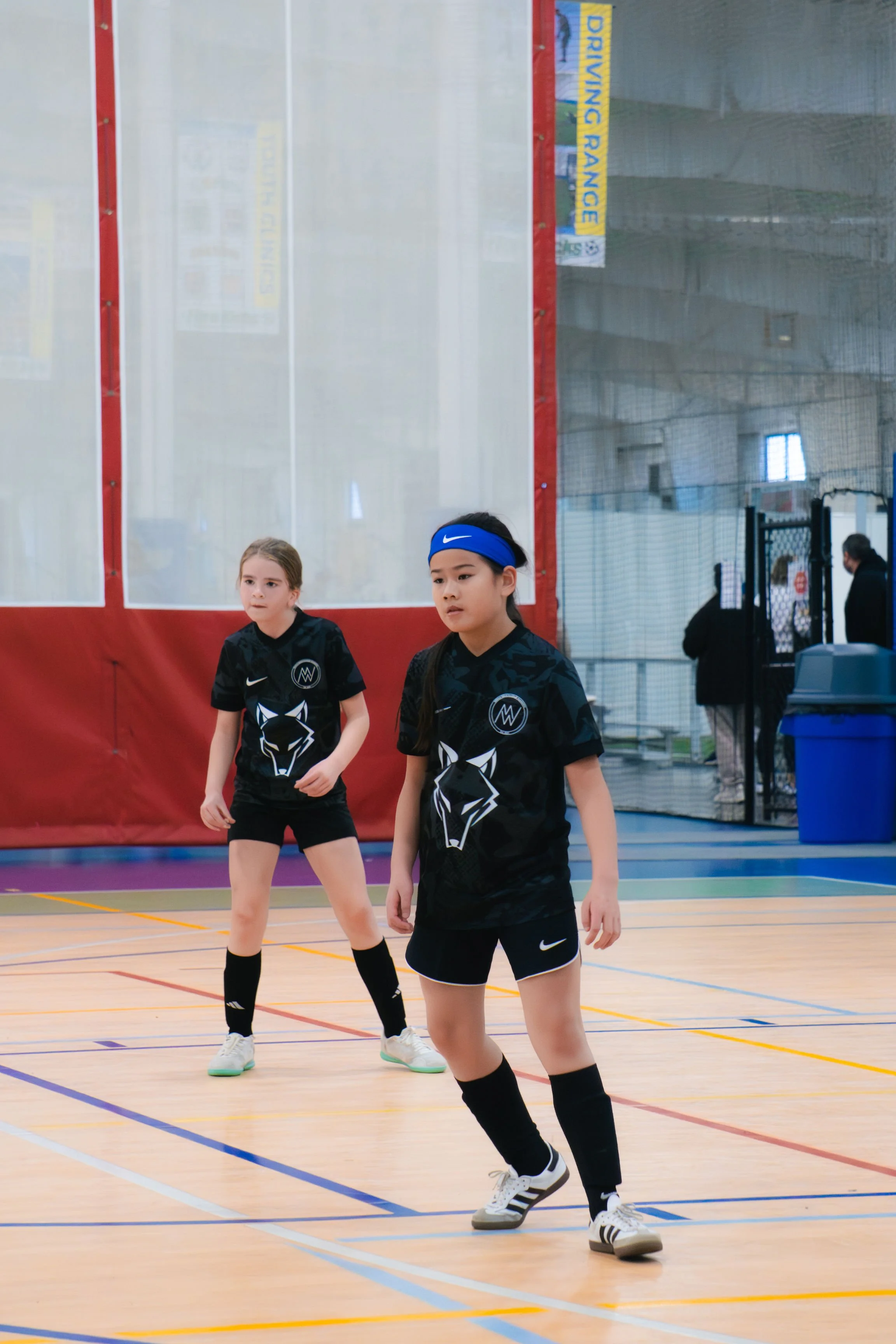 Two young girls playing indoor soccer or futsal on a court, wearing black soccer jerseys with wolf logos, black shorts, and black knee-high socks.