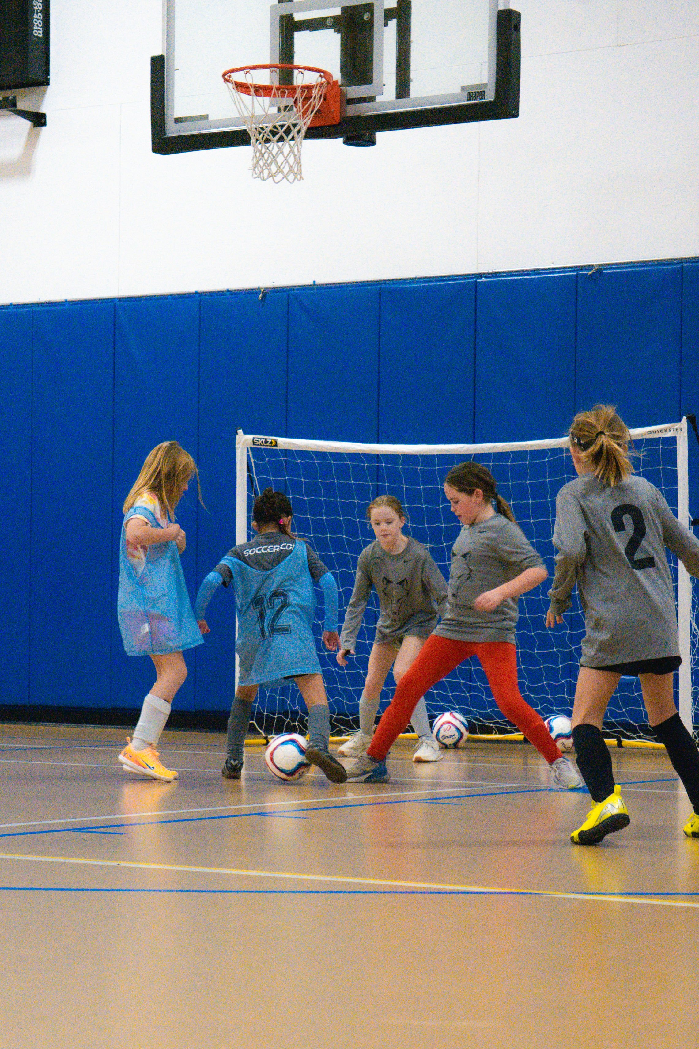 Children playing indoor soccer in a gym, with a goal and basketball hoop visible.