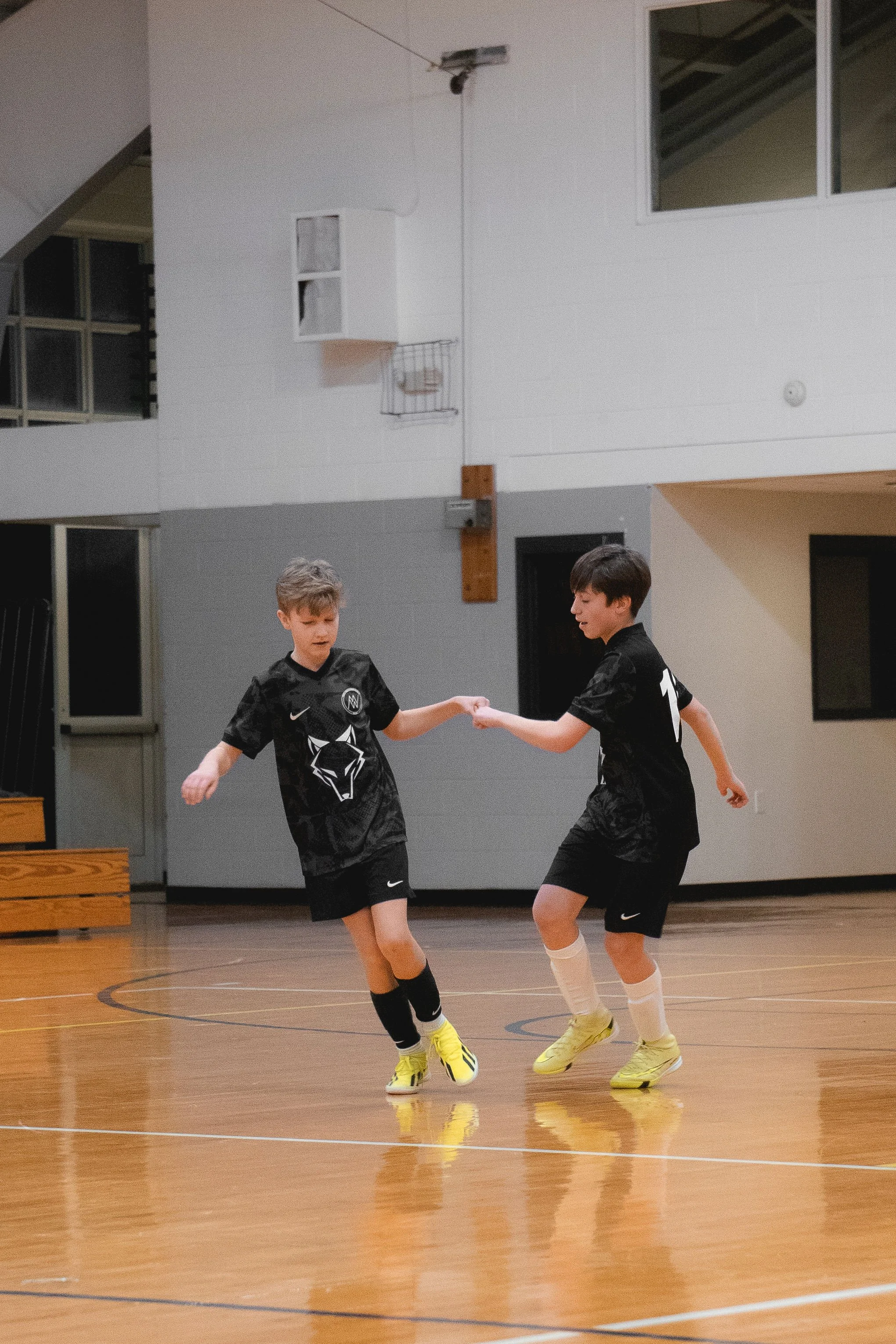Two young boys playing indoor soccer on a wooden court, holding hands, wearing black jerseys and yellow shoes.
