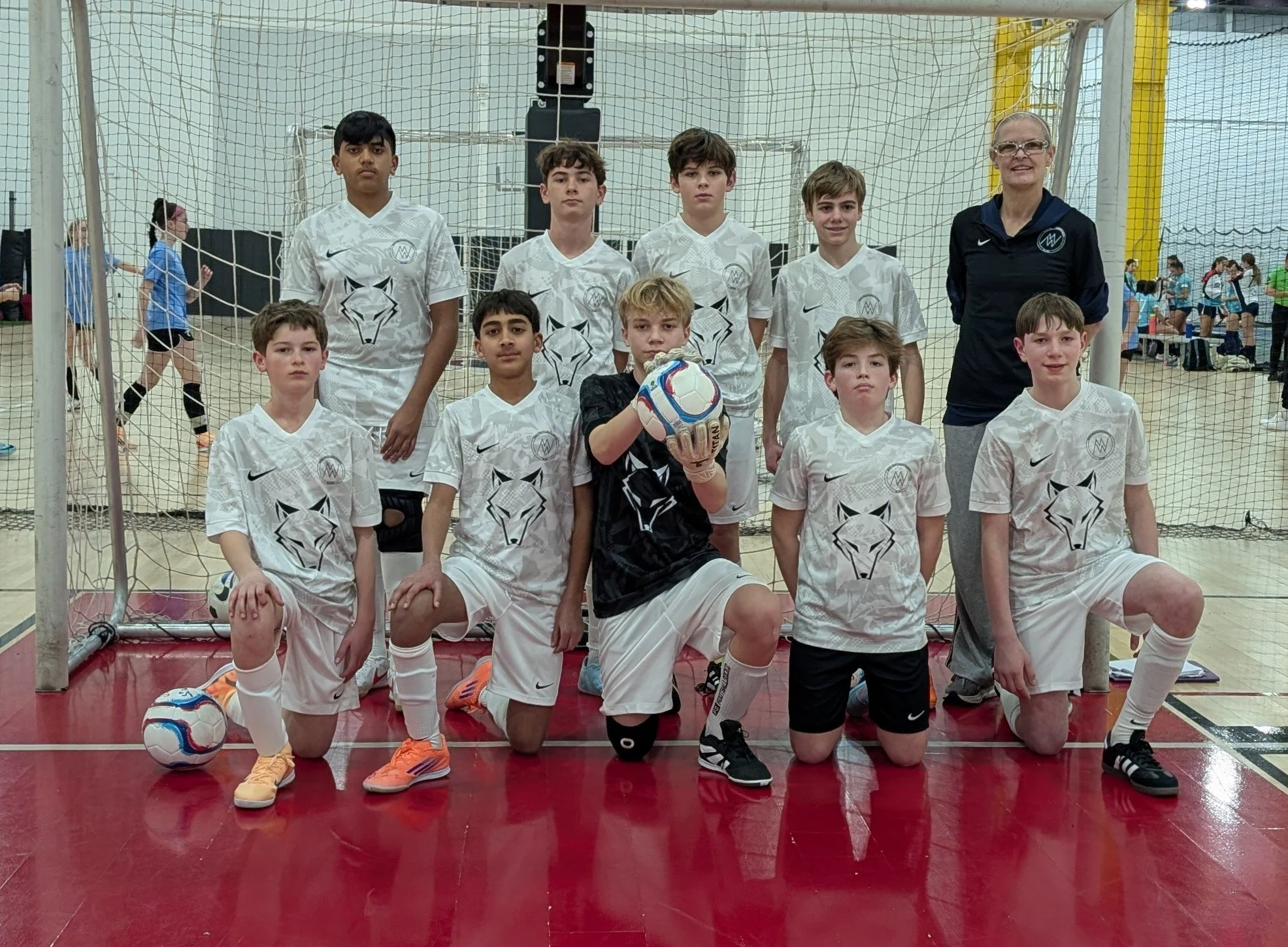 A youth soccer team posing indoors in front of a goal, wearing matching white uniforms with a wolf logo, with a school or sports facility in the background.