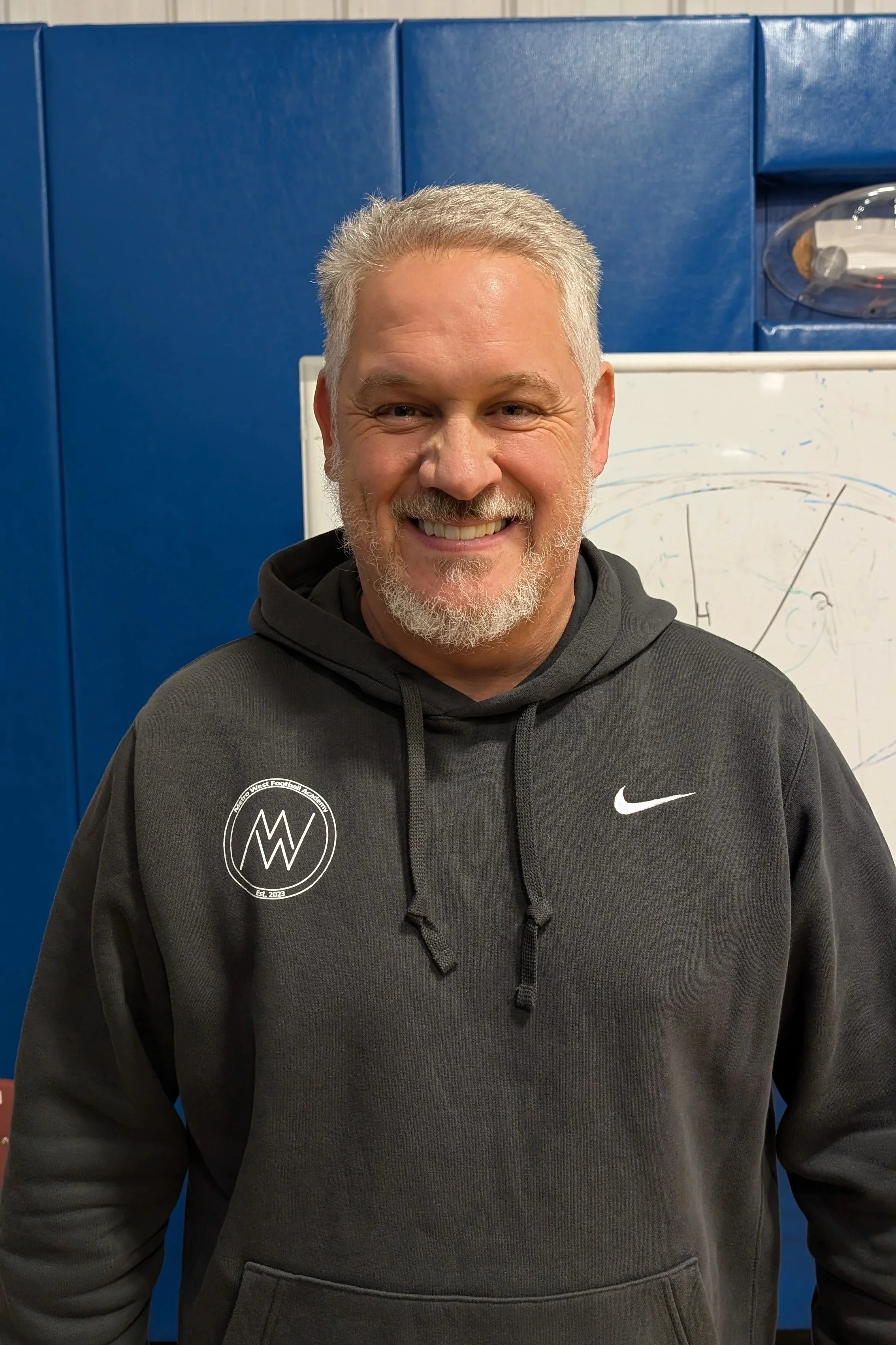 A smiling middle-aged man with gray hair and beard, wearing a gray Nike hoodie with a logo on the chest, standing in front of a whiteboard with a diagram, in a room with blue padded walls.