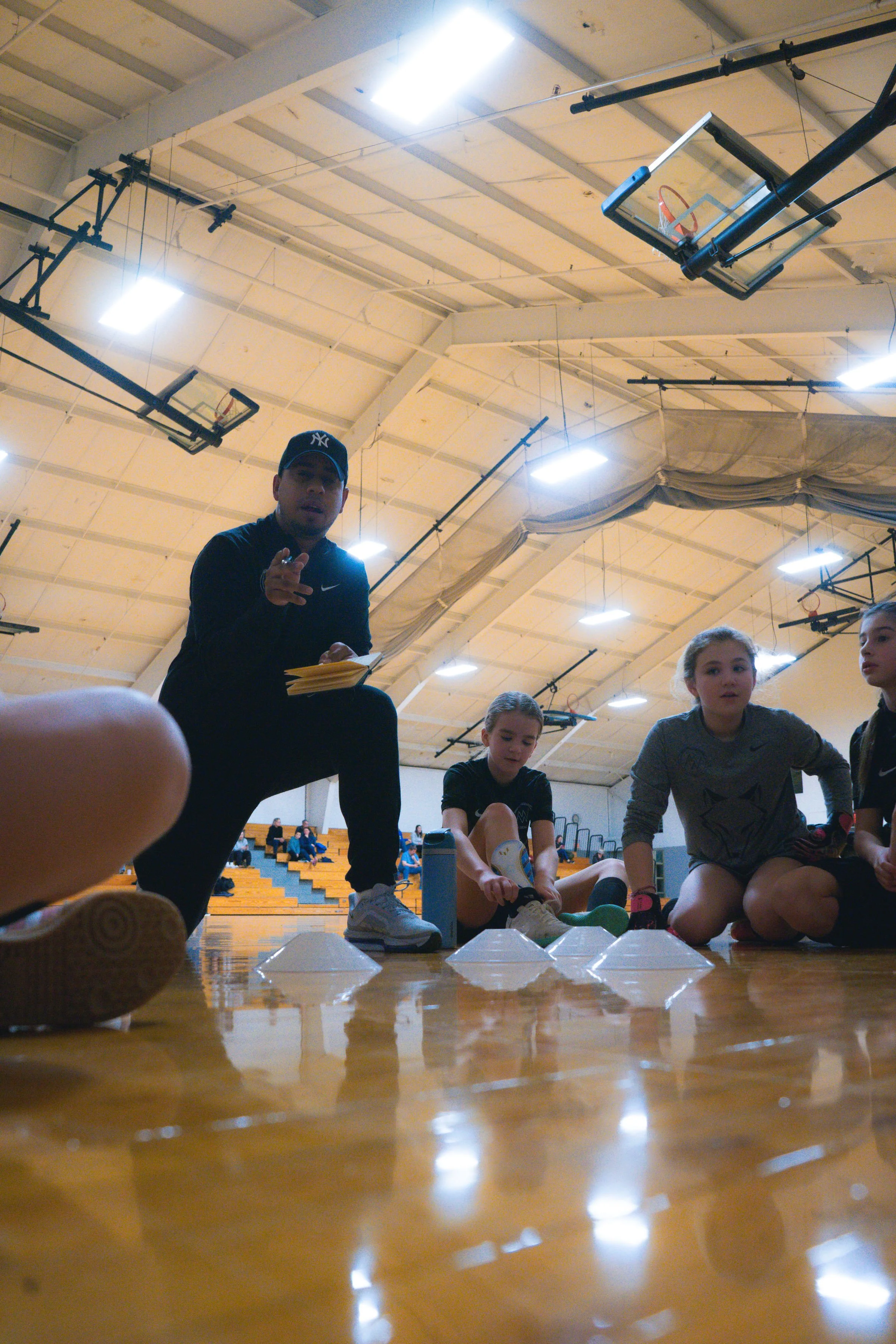 A coach or instructor with a notebook talking to young girls seated on a gymnasium floor during a sports practice or class.