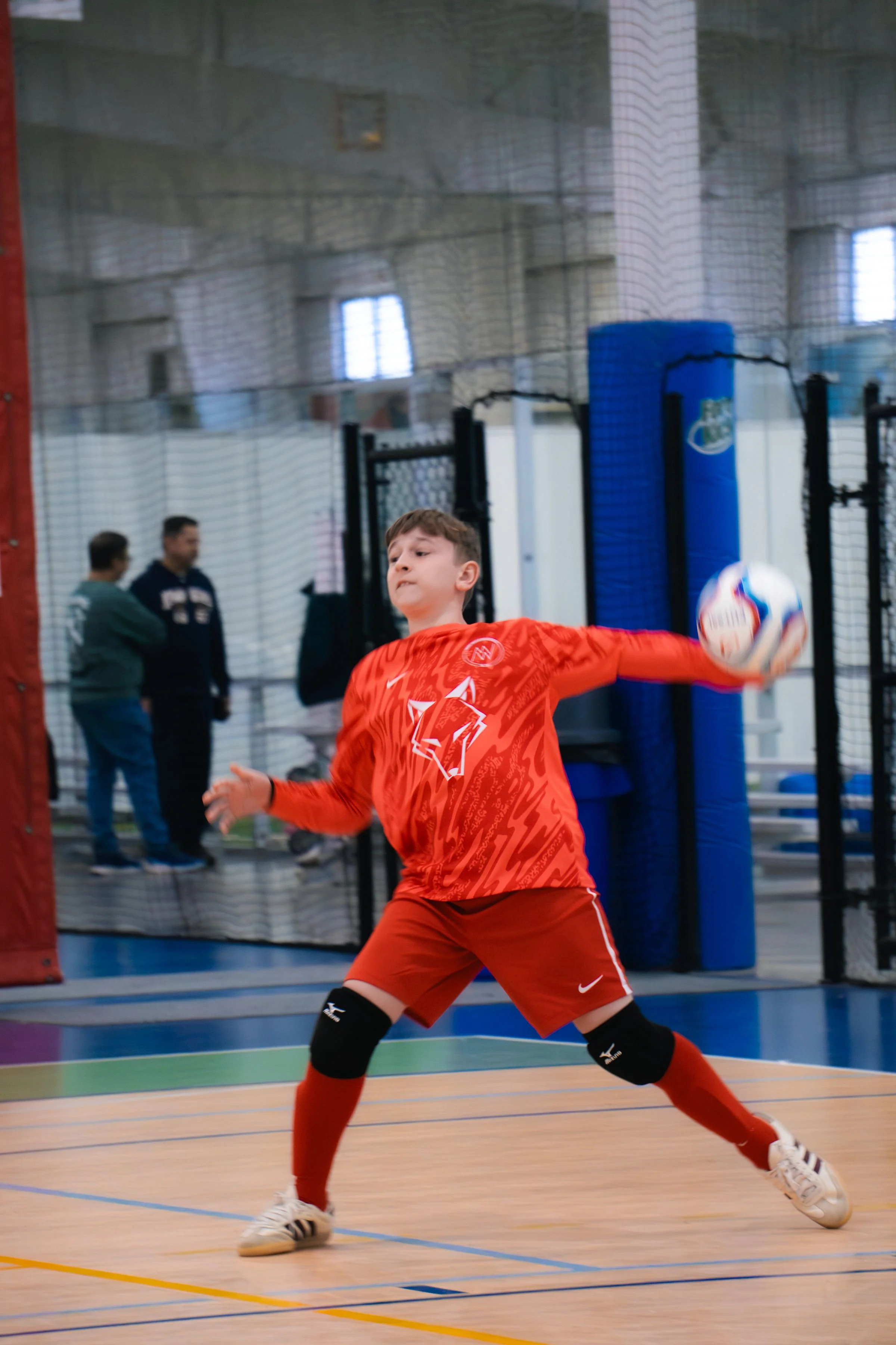 Young boy in red sports uniform is playing indoor soccer or futsal, kicking the ball with his right leg. In the background, three adults are observing behind a net, in an indoor sports facility.
