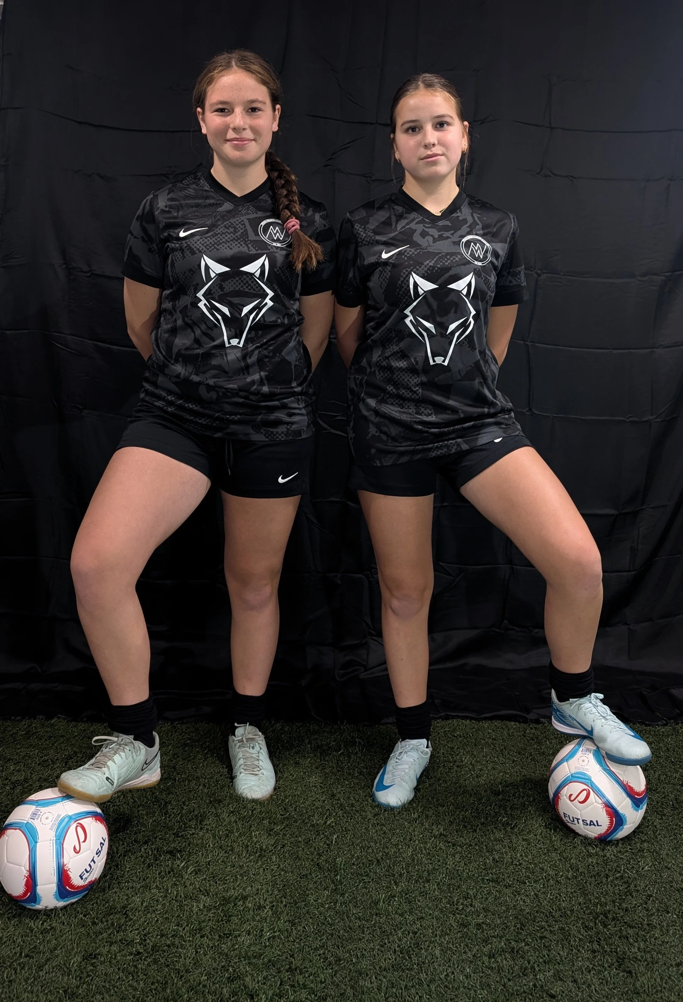 Two young female soccer players standing side by side on a green turf with a black backdrop. They wear matching black jerseys with wolf logos, black shorts, and soccer cleats, each with a ball at their feet.