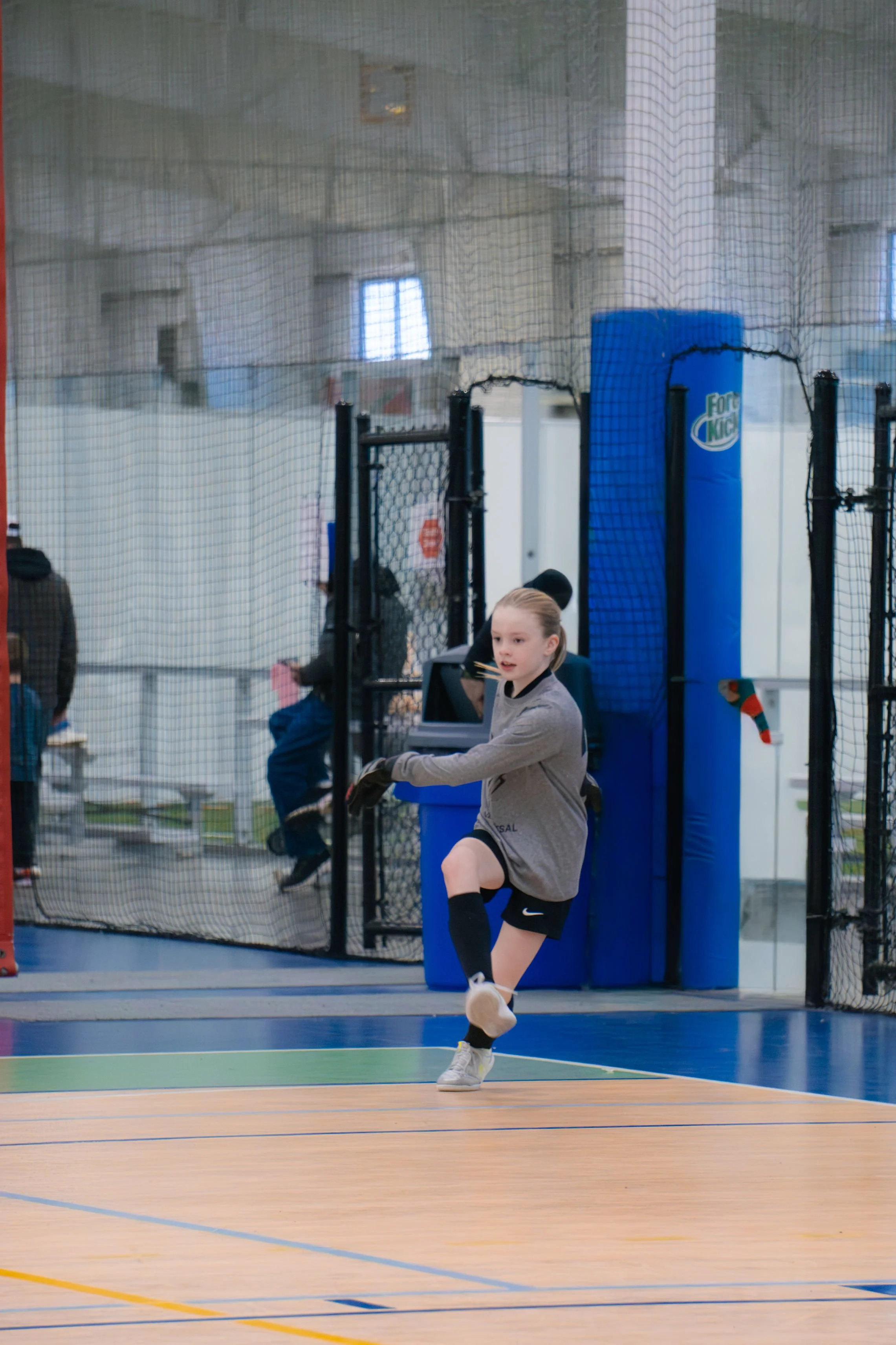 A young girl in athletic clothing is stretching her leg in an indoor sports facility with a basketball court, surrounded by a safety net and padded columns.