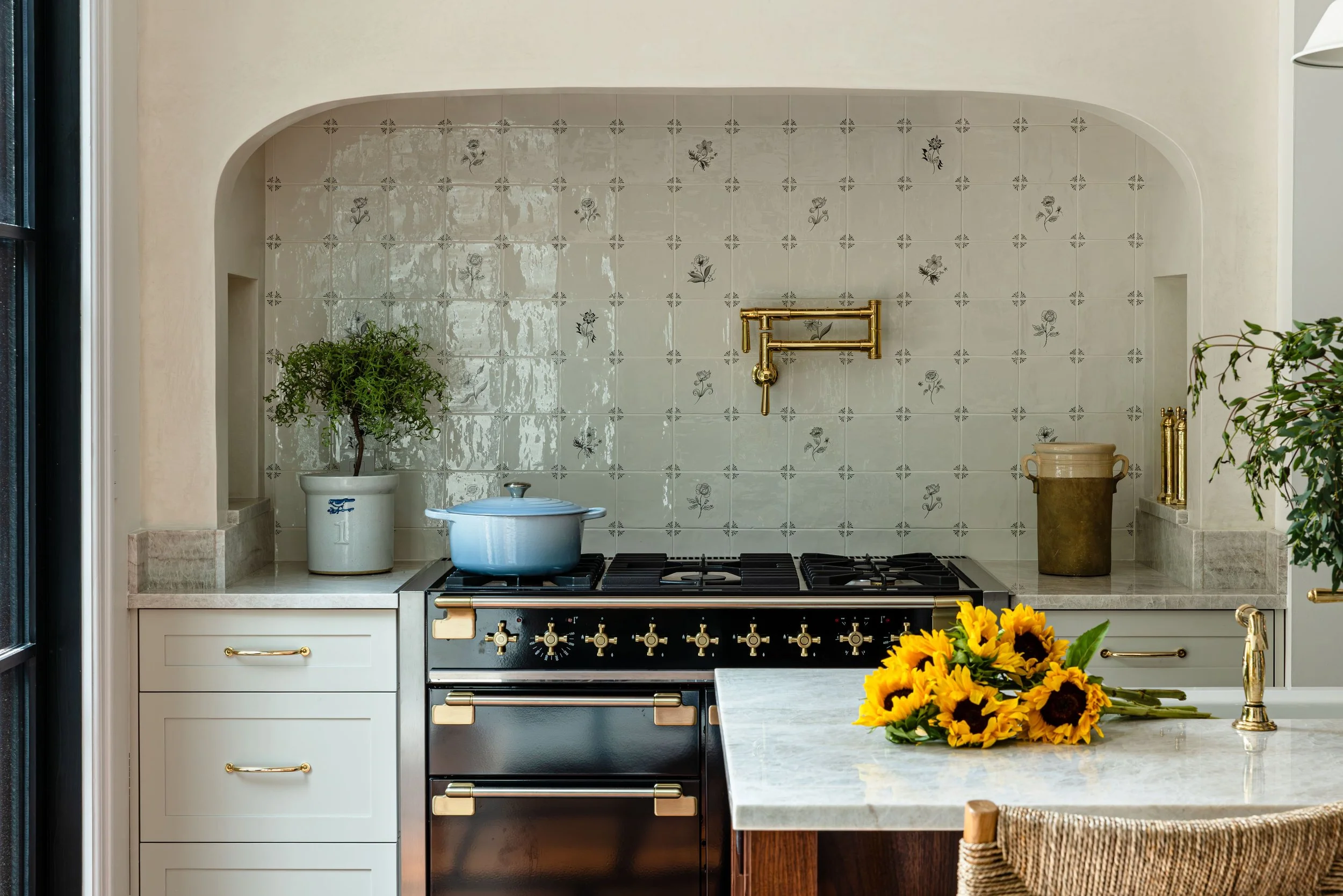 Kitchen with a white counter, gold fixtures, a blue pot on a black and gold stove, a potted plant, a beige container, and a bouquet of sunflowers on the counter.