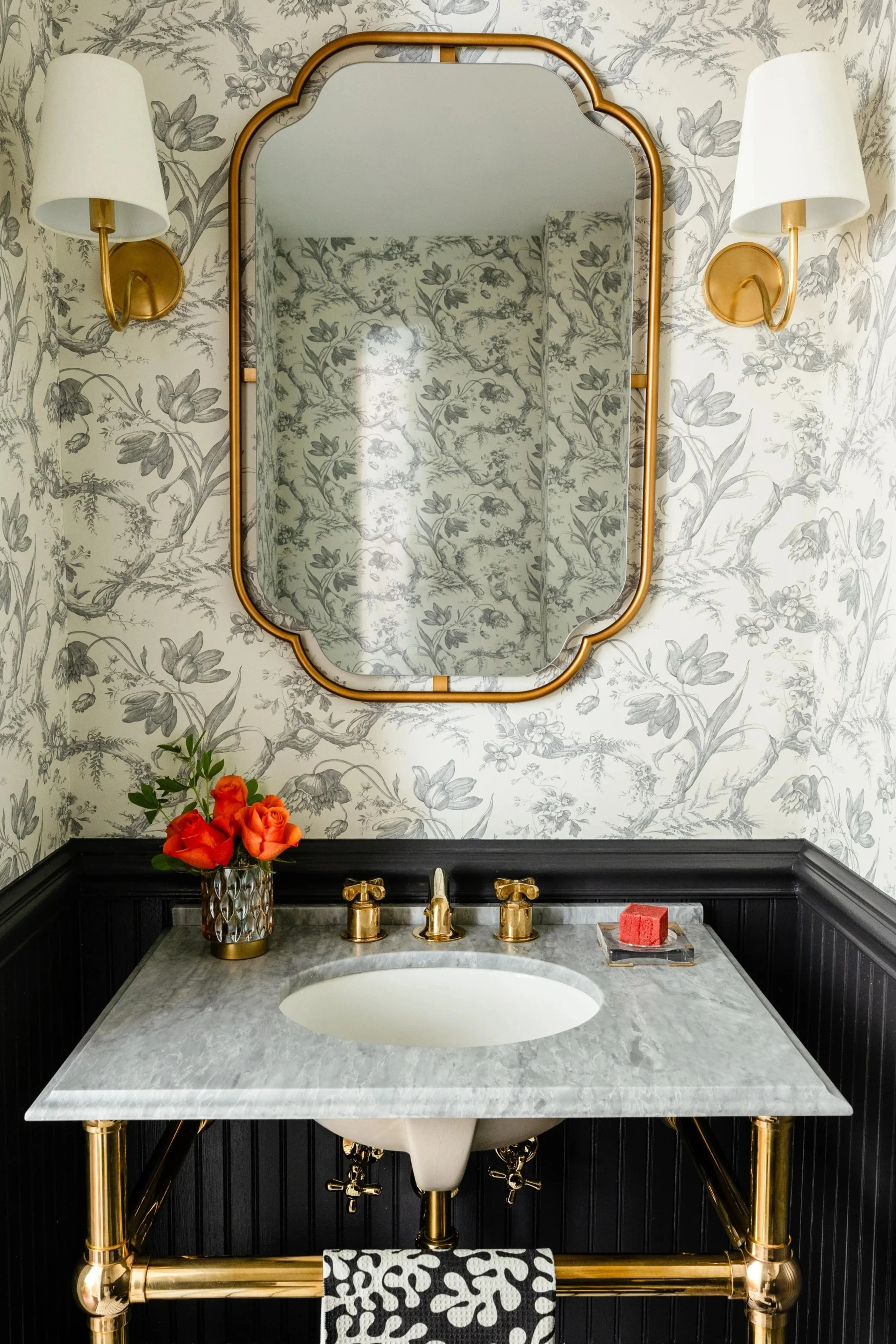 Bathroom vanity with marble countertop, gold fixtures, a mirror, wall sconces, a vase with orange roses, and a soap on a tray.