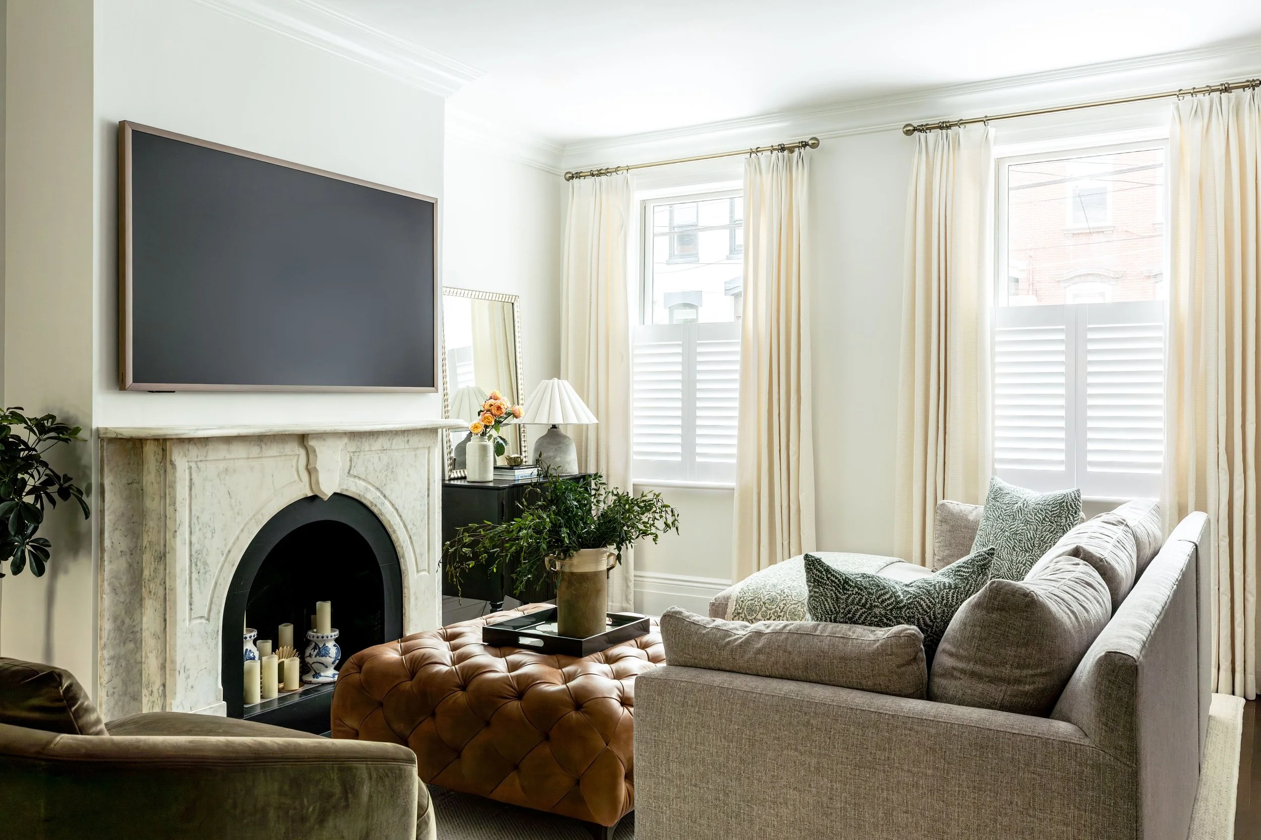 Living room with a fireplace, a wall-mounted TV, a marble fireplace mantel, beige curtains, a gray sofa with patterned pillows, a tufted leather ottoman, a side table with a lamp, flowers, and a mirror.