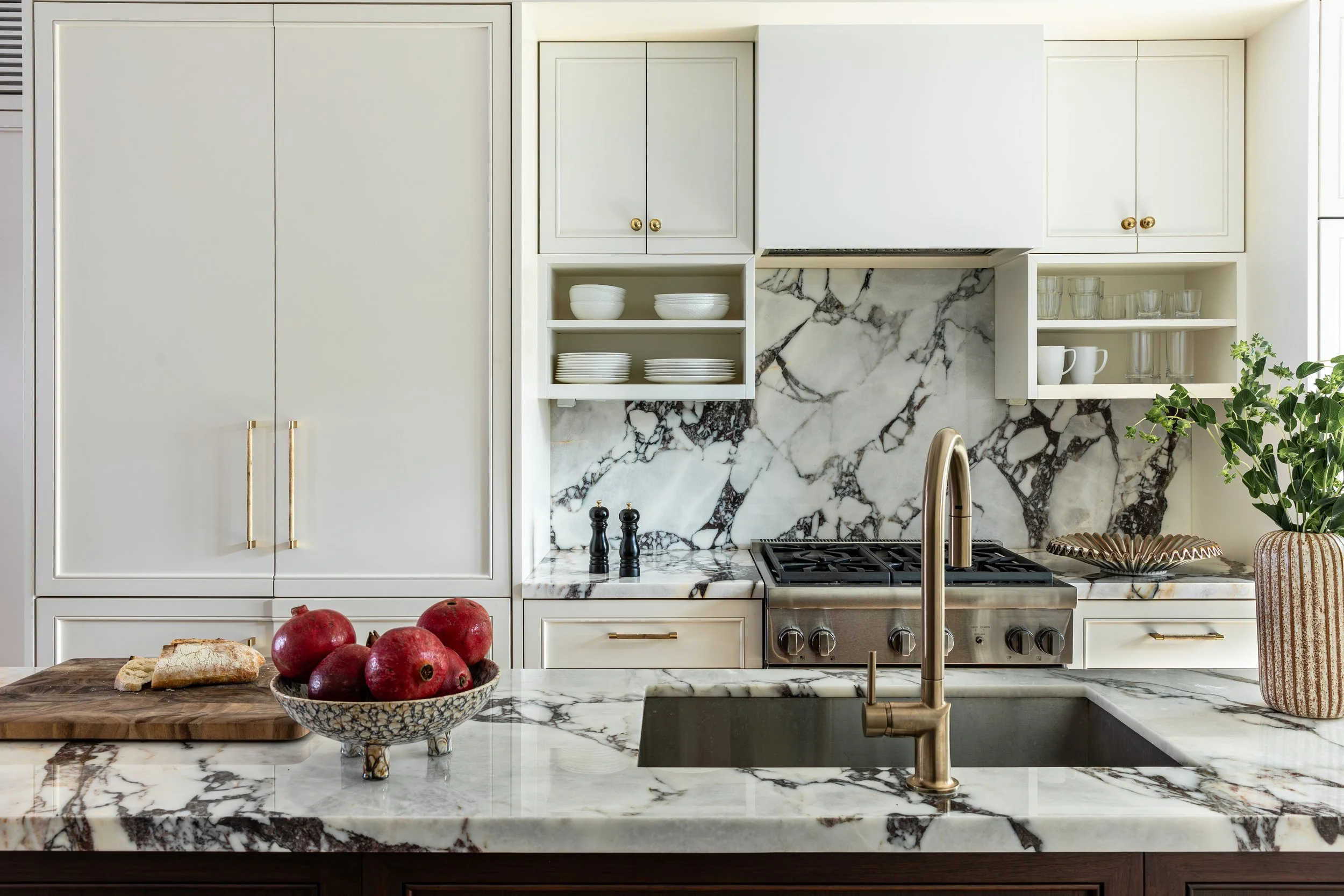 Kitchen with white cabinets, marble backsplash, and a marble countertop island. Pomegranate bowl, bread, black salt and pepper shakers, kitchen sink with a gold faucet, stove, plant, and open shelves with dishes.