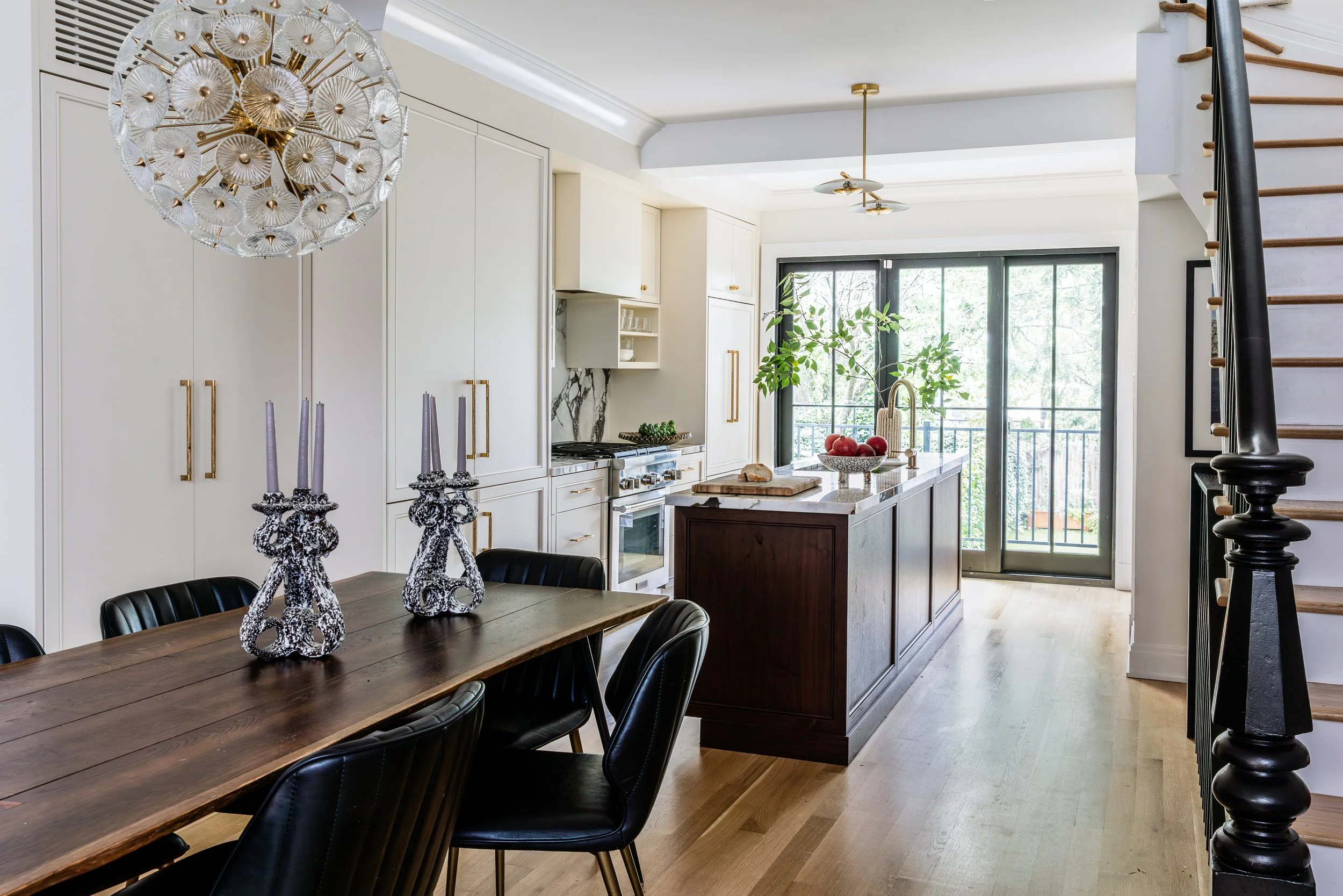 A modern kitchen with white cabinetry, a dark wood kitchen island, and a large window with trees outside. There is a dining table with black chairs and decorative candle holders, and a staircase is visible on the right.