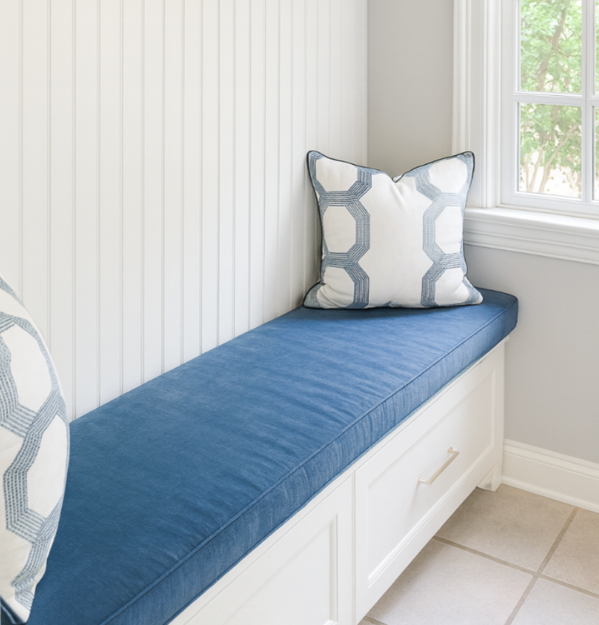 White window seat with a blue cushion and white pillows with blue geometric patterns, located by a window with a view of green trees outside.