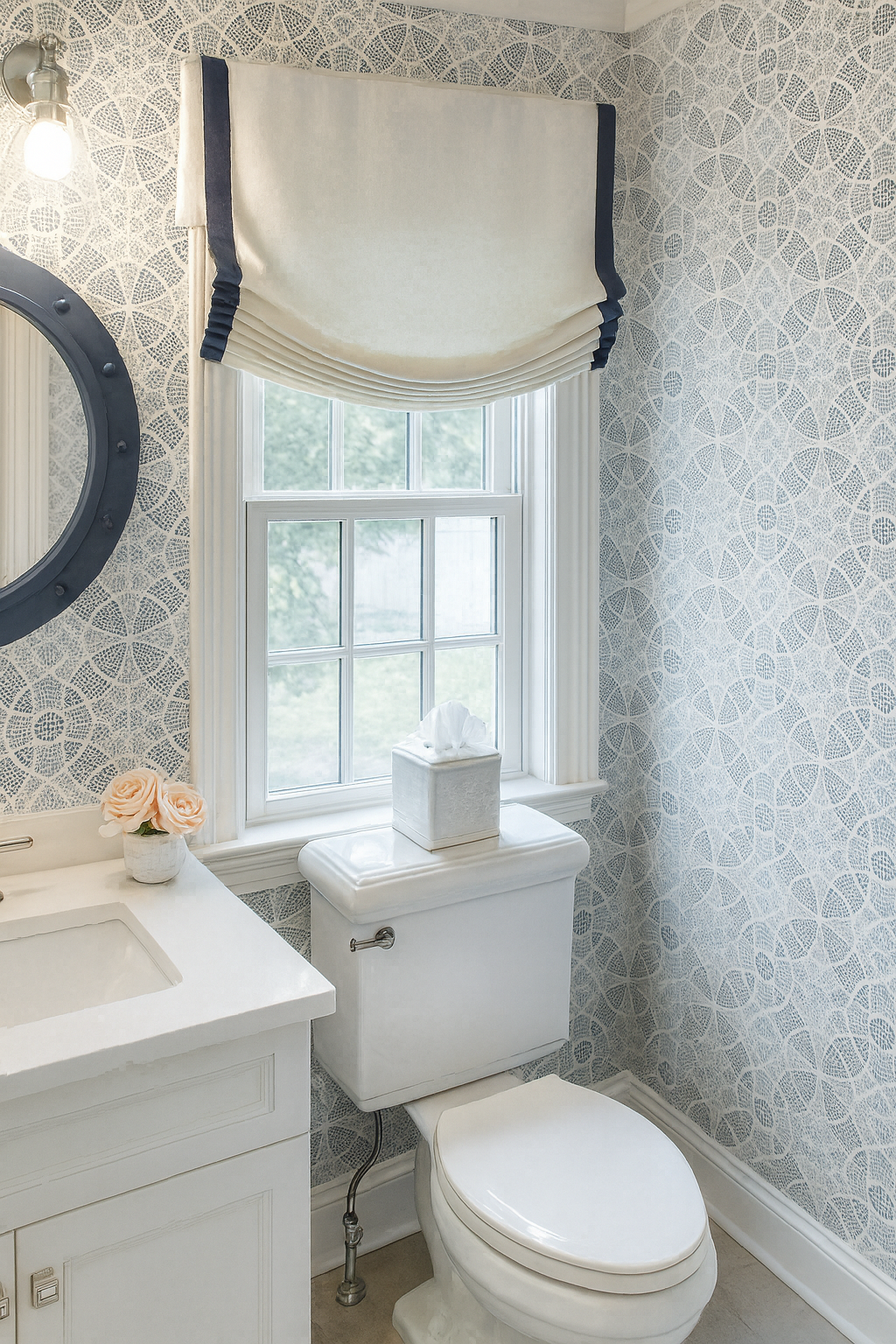 Bathroom with blue and white patterned wallpaper, white vanity with a small flower vase, tissue box on the toilet tank, window with draped valance, and a round mirror with bulb lighting.