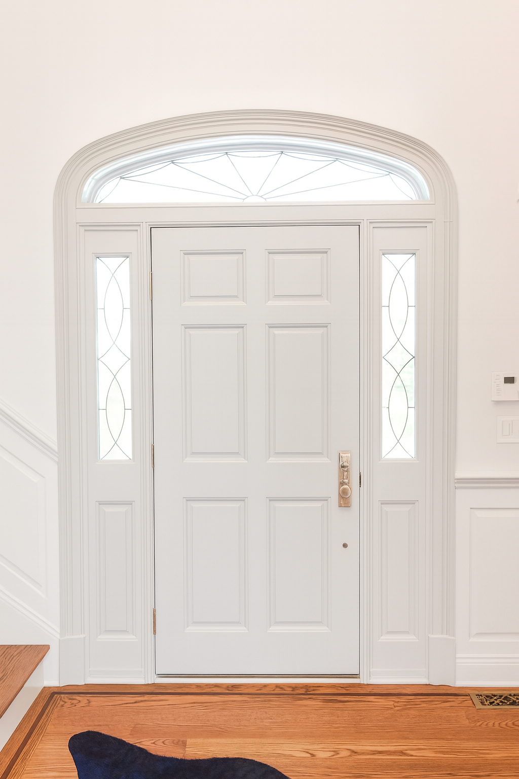 White front door with glass side panels and transom window, with hardwood floor and a dark doormat.