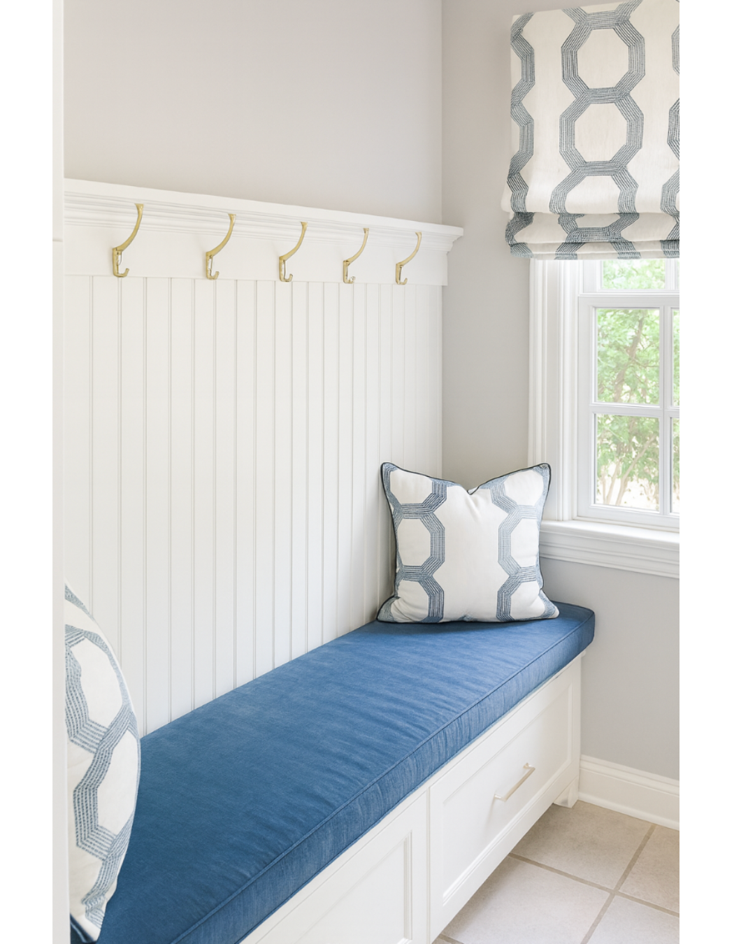 Window seat with blue cushion and white pillows with blue geometric patterns, white beadboard wall with gold hooks, window with a patterned Roman shade, and tiled floor.