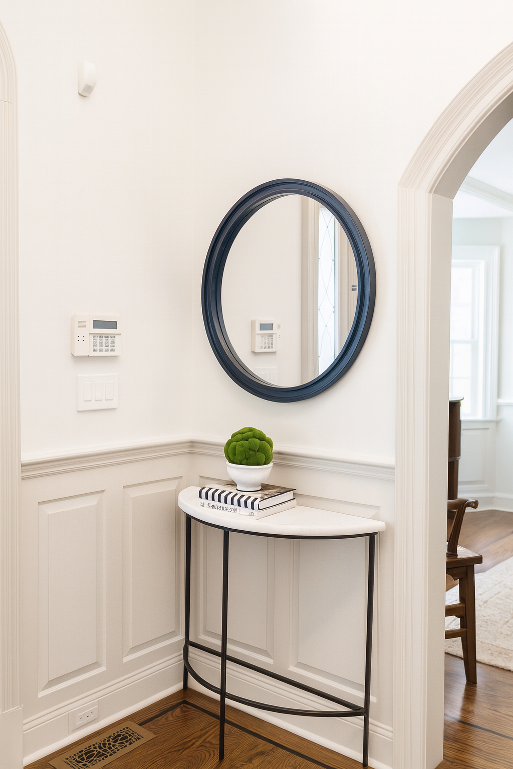 Entryway with a white wall, black oval mirror, small white console table with a green moss ball plant and books, and a wooden chair visible through a doorway.