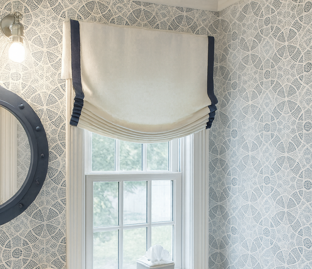Interior view of a room corner with a window featuring a beige and navy blue window treatment, patterned wallpaper, and a round mirror to the left.