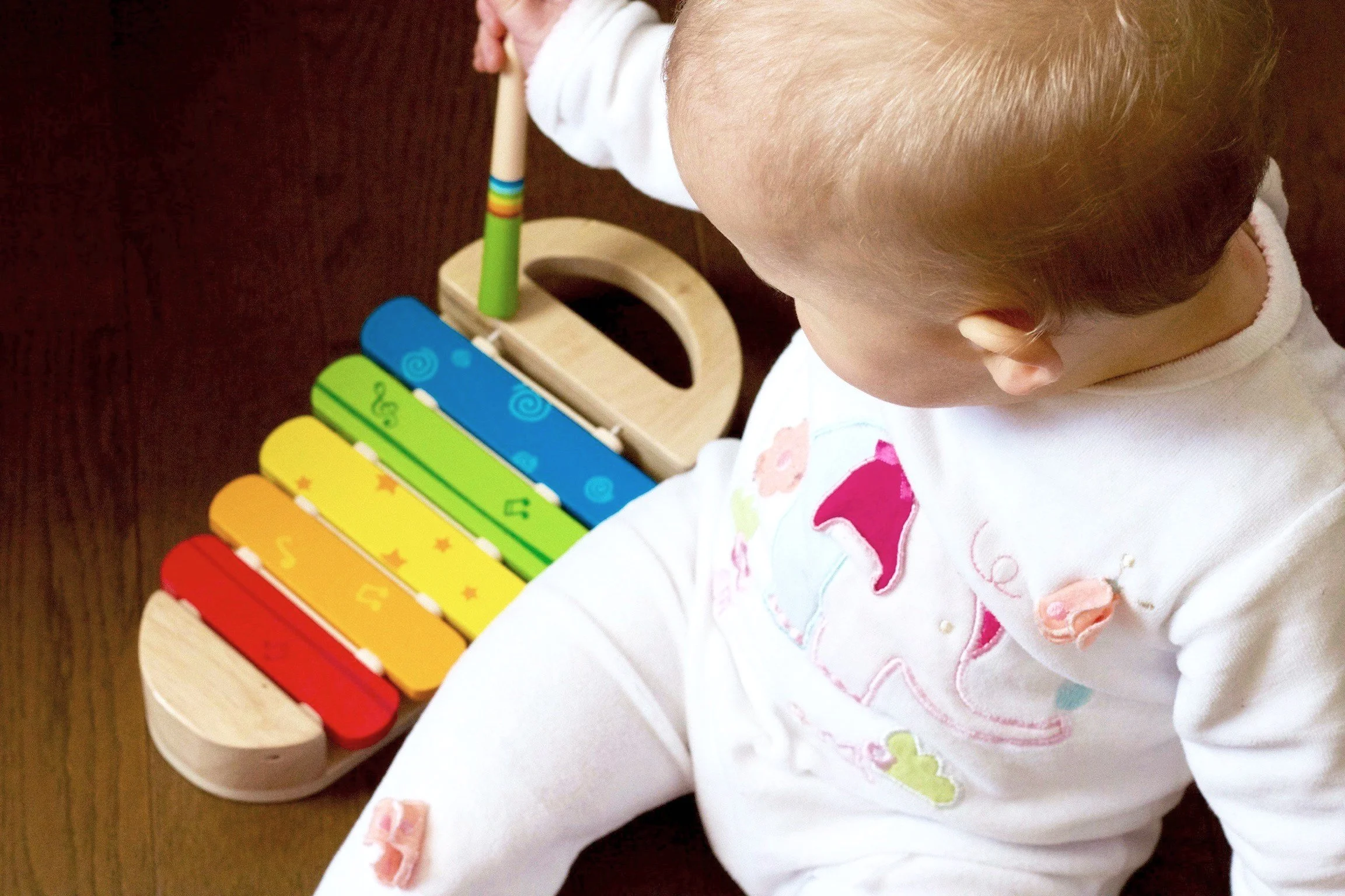 A young child with light brown or blonde hair wearing a white shirt sitting on a wooden floor, playing a colorful xylophone.