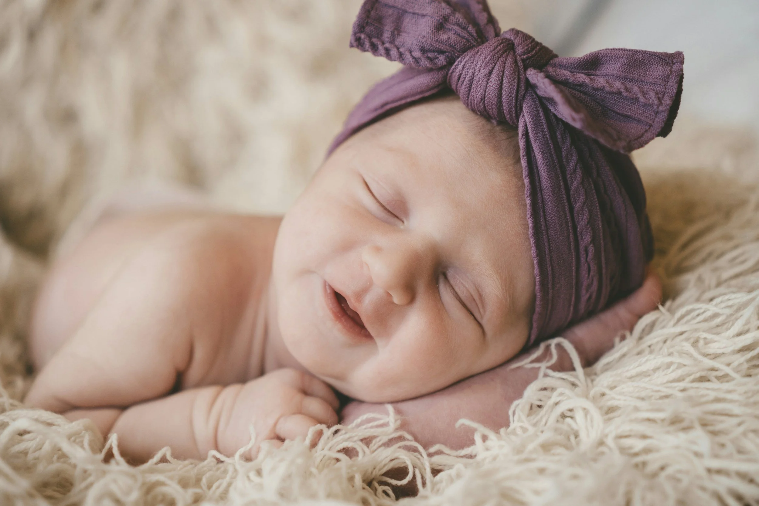 A smiling sleeping baby wearing a purple headband with a bow, lying on a fluffy cream-colored blanket.
