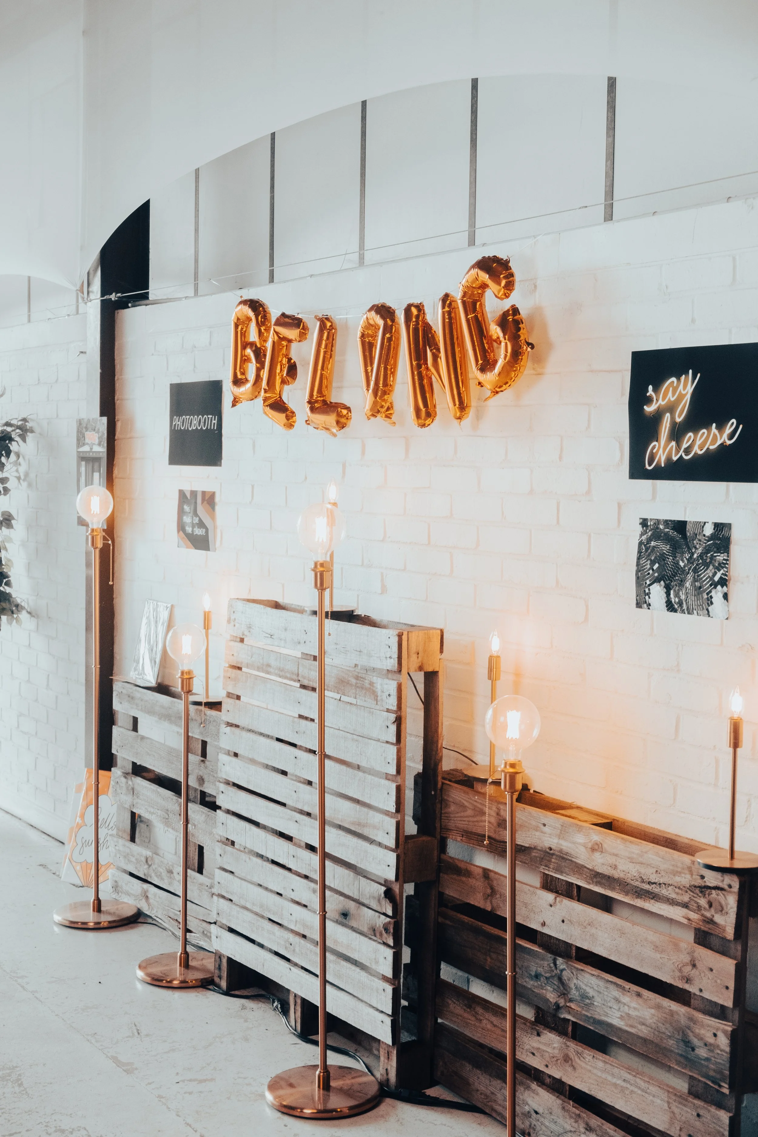 Decorative setup with gold balloon letters spelling 'BELLY', four copper floor lamps with exposed bulbs, white brick wall, and various signage including 'Photobooth' and 'say cheese' for a celebration event.