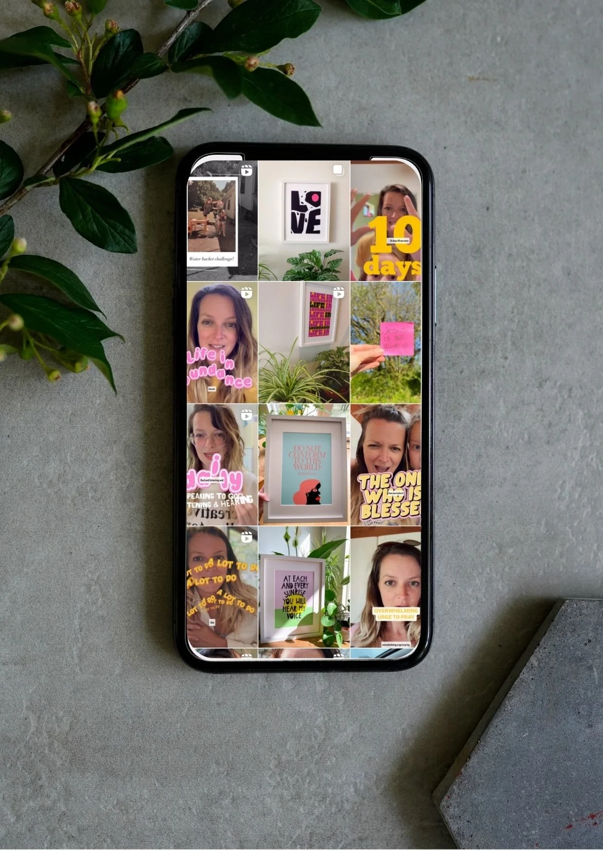 Smartphone displaying a social media grid with photos, quotes, and stickers, placed on a concrete surface near a green leafy plant and a stone block.