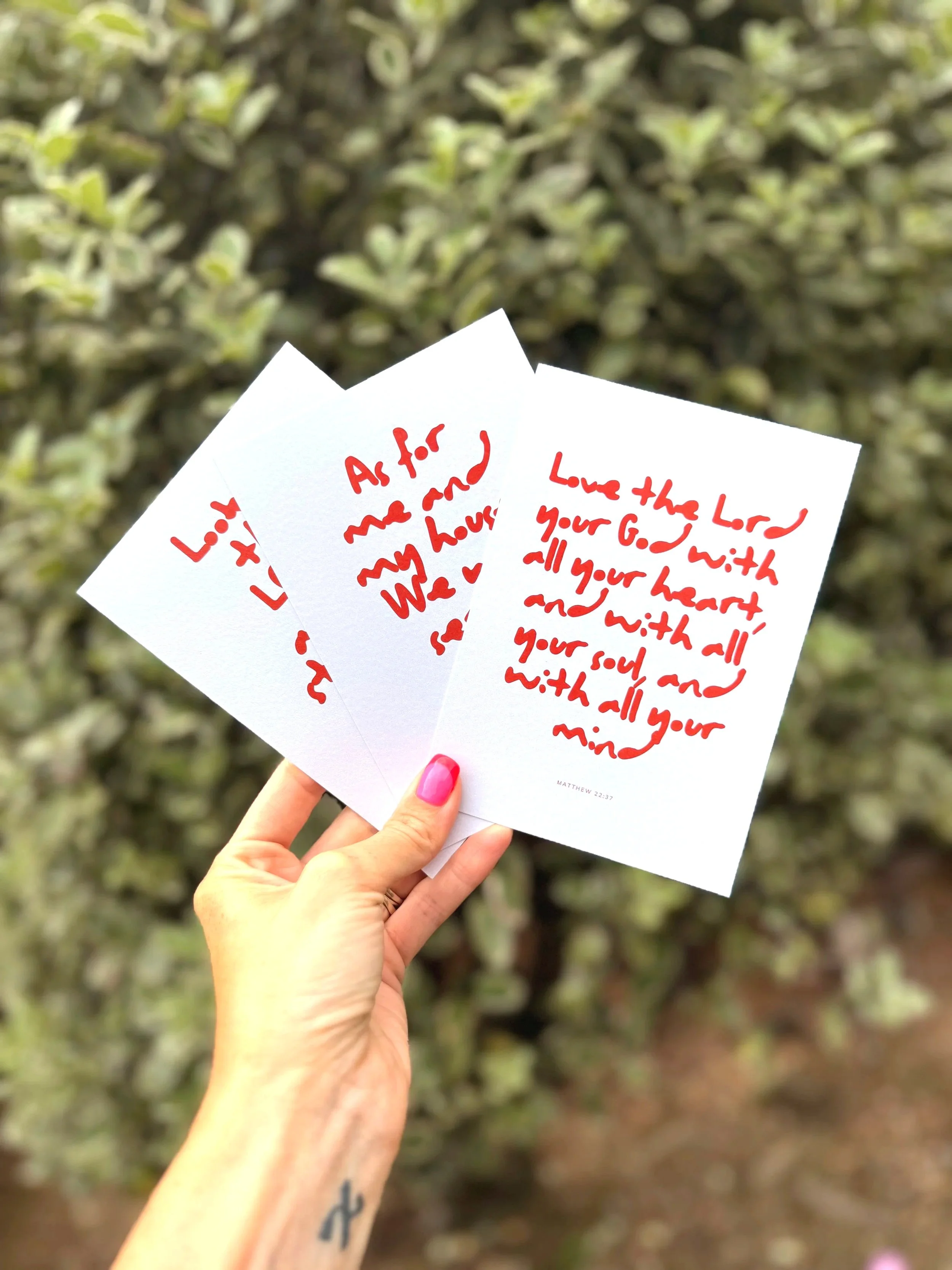 Hand with pink nail polish holding three cards with red handwritten text in front of a leafy background.