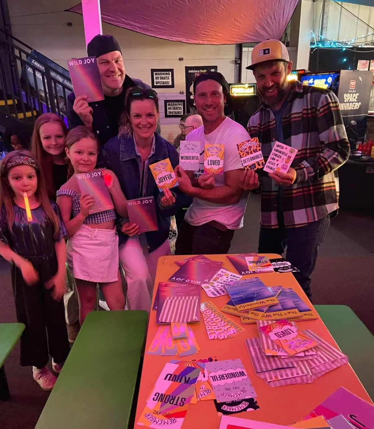 Group of people, including children and adults, gathered around a table at a festive event, holding colorful cards with words like 'Wild Joy,' 'Joyful,' 'Loved,' and 'Wonderful.'