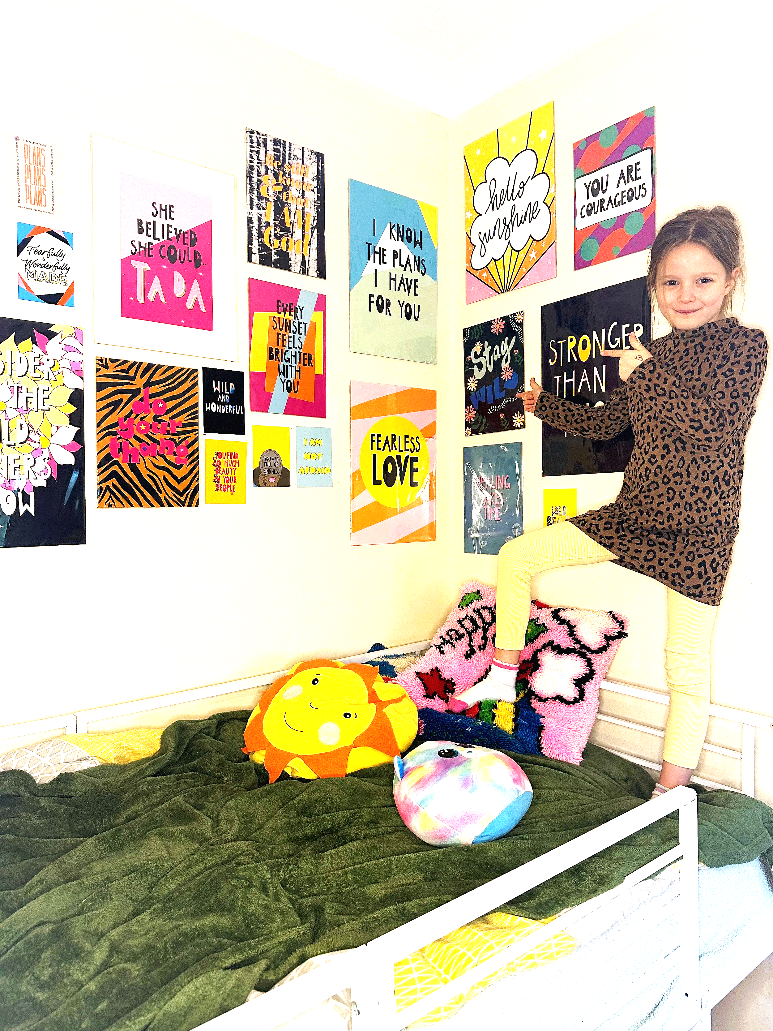 A girl with brown hair in a braid, wearing a leopard print top and yellow pants, sitting on a bed covered with black and pink blankets. She is pointing at a sign on the wall that says 'Stronger Than' and is smiling. The wall behind her is decorated with colorful posters with inspirational messages. There are plush toys and a pink rug on the bed.