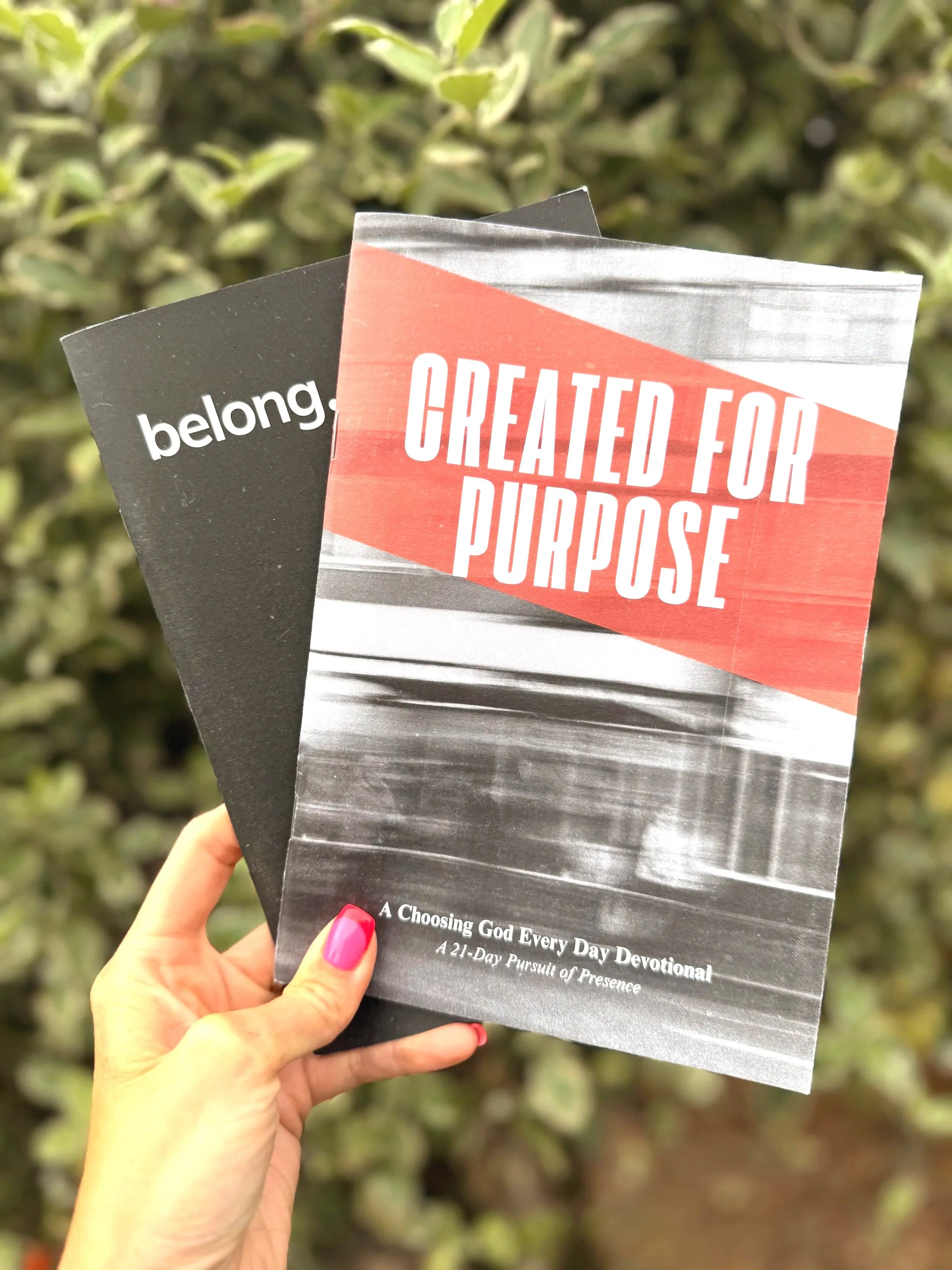 A hand holding two devotional books titled 'Created for Purpose' and 'belong' against a background of green foliage.