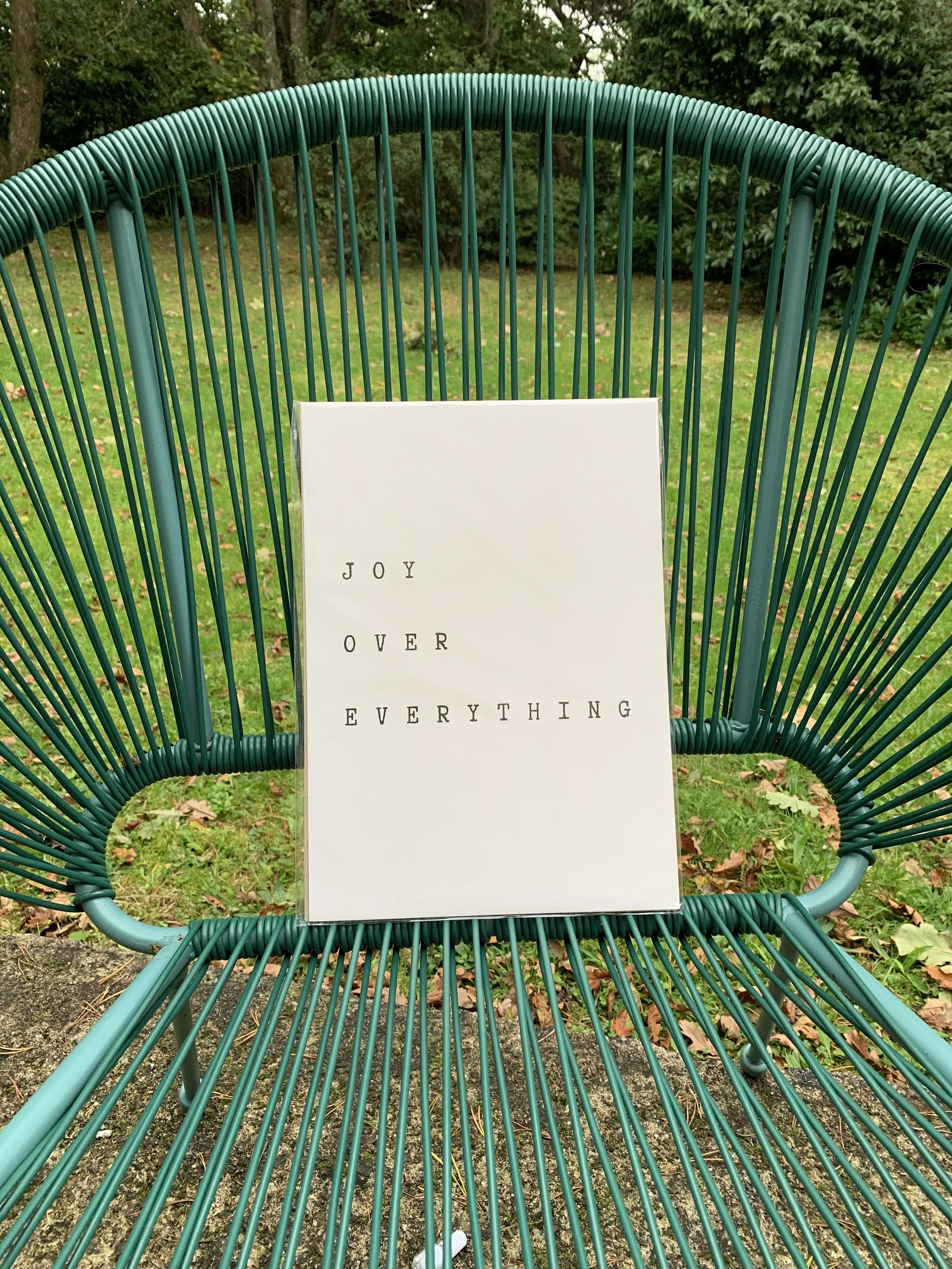 A white card with the words 'Joy Over Everything' in black text, placed on a green metal wire chair outdoors on grass with fallen leaves and green trees in the background.