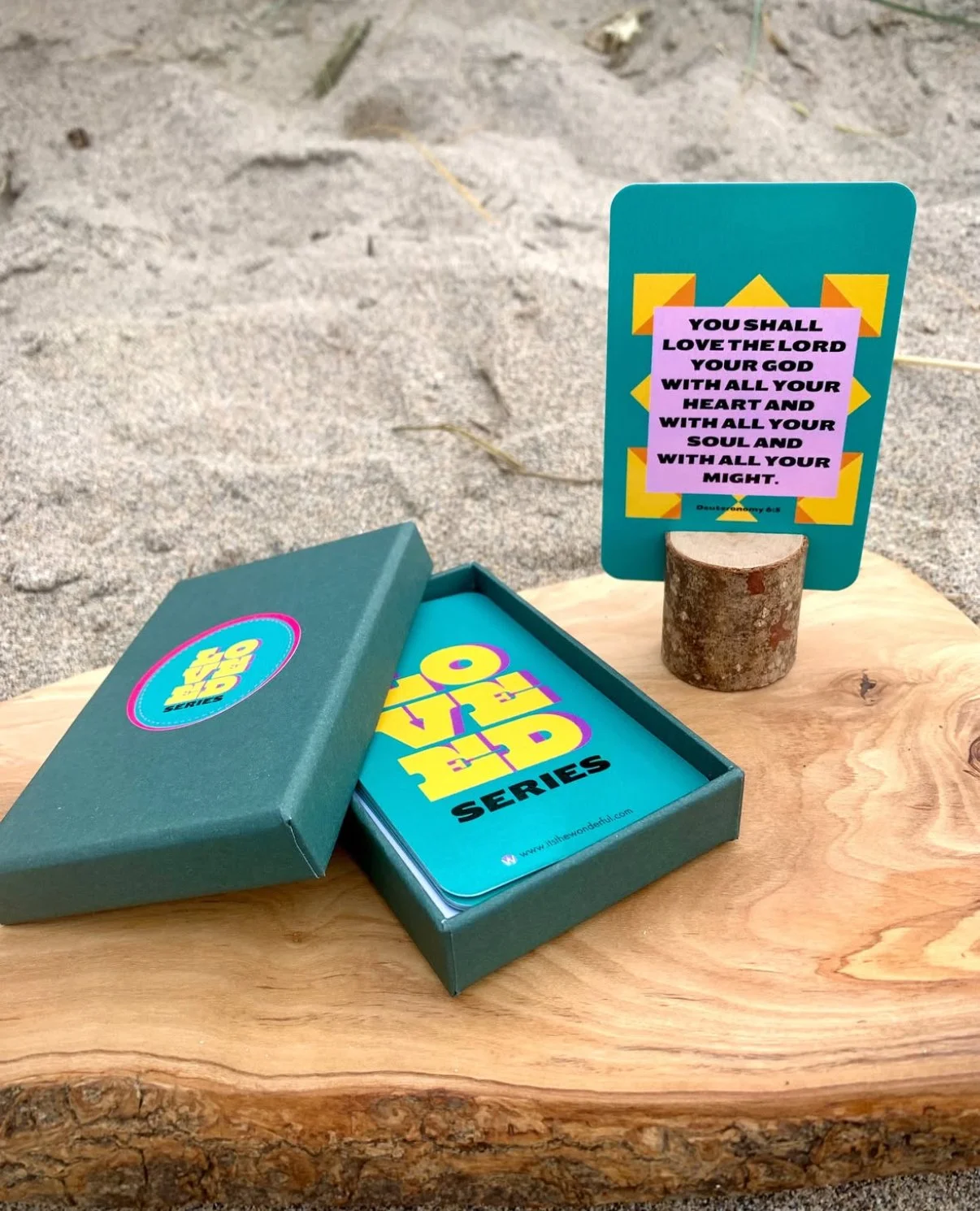A card game called 'Safe to Say' in a green box on a wooden surface, with a colorful scripture card standing on a small log, displaying a Bible verse.