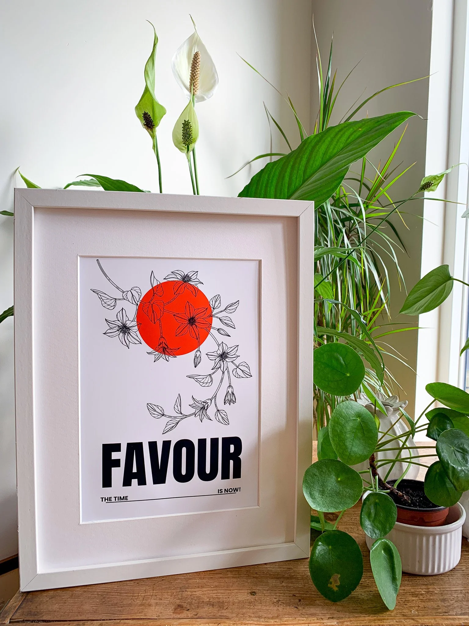 A framed poster with a red circle, black line drawing of flowers, and the word FAVOUR in bold black letters on a white background, surrounded by green houseplants near a window.