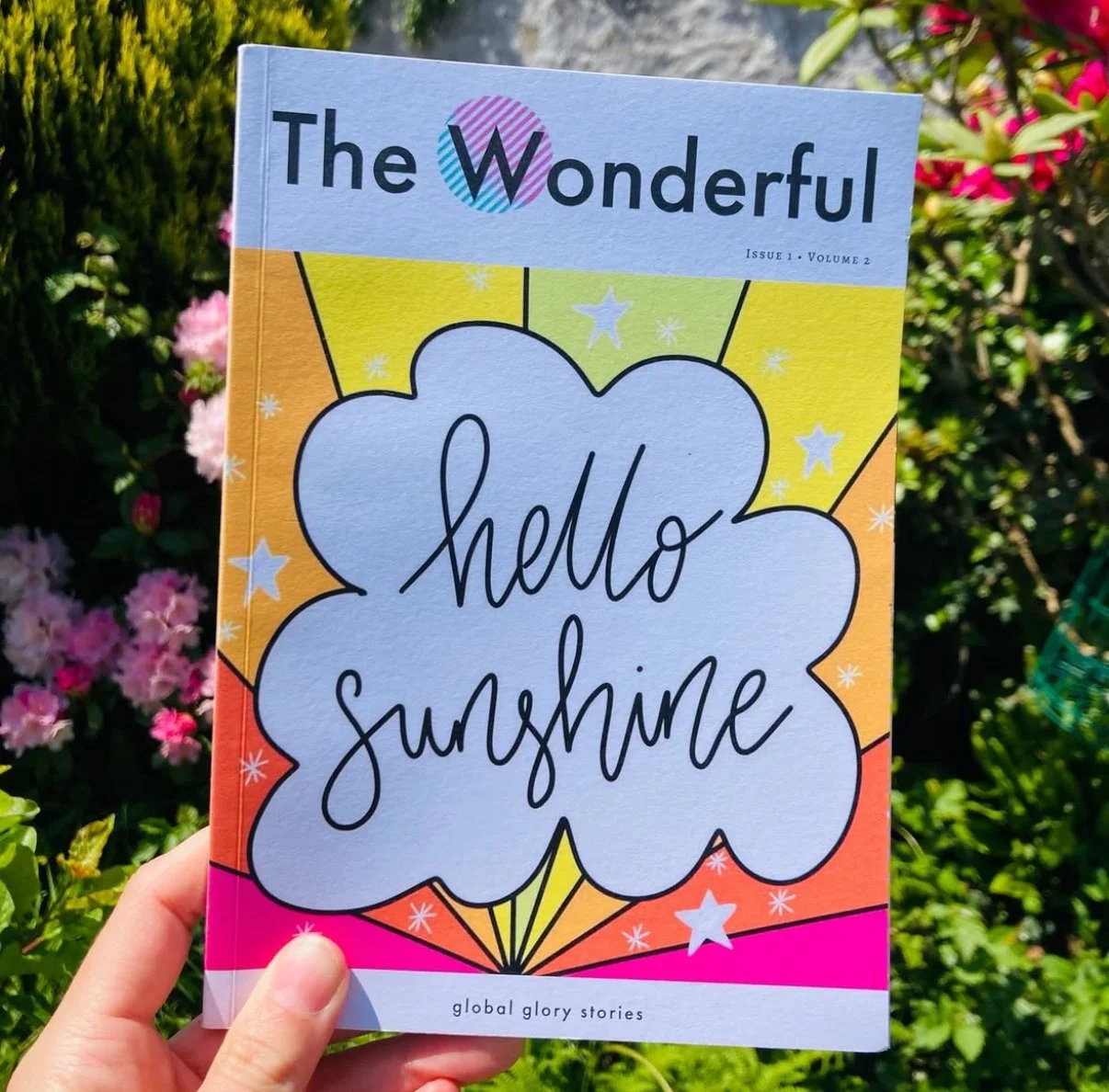 A colorful magazine titled "The Wonderful" with a large cloud-shaped speech bubble that says "hello sunshine." The background features bright yellow, orange, and pink sections with stars and sparkles, with a hand holding it outdoors against green foliage and pink flowers.